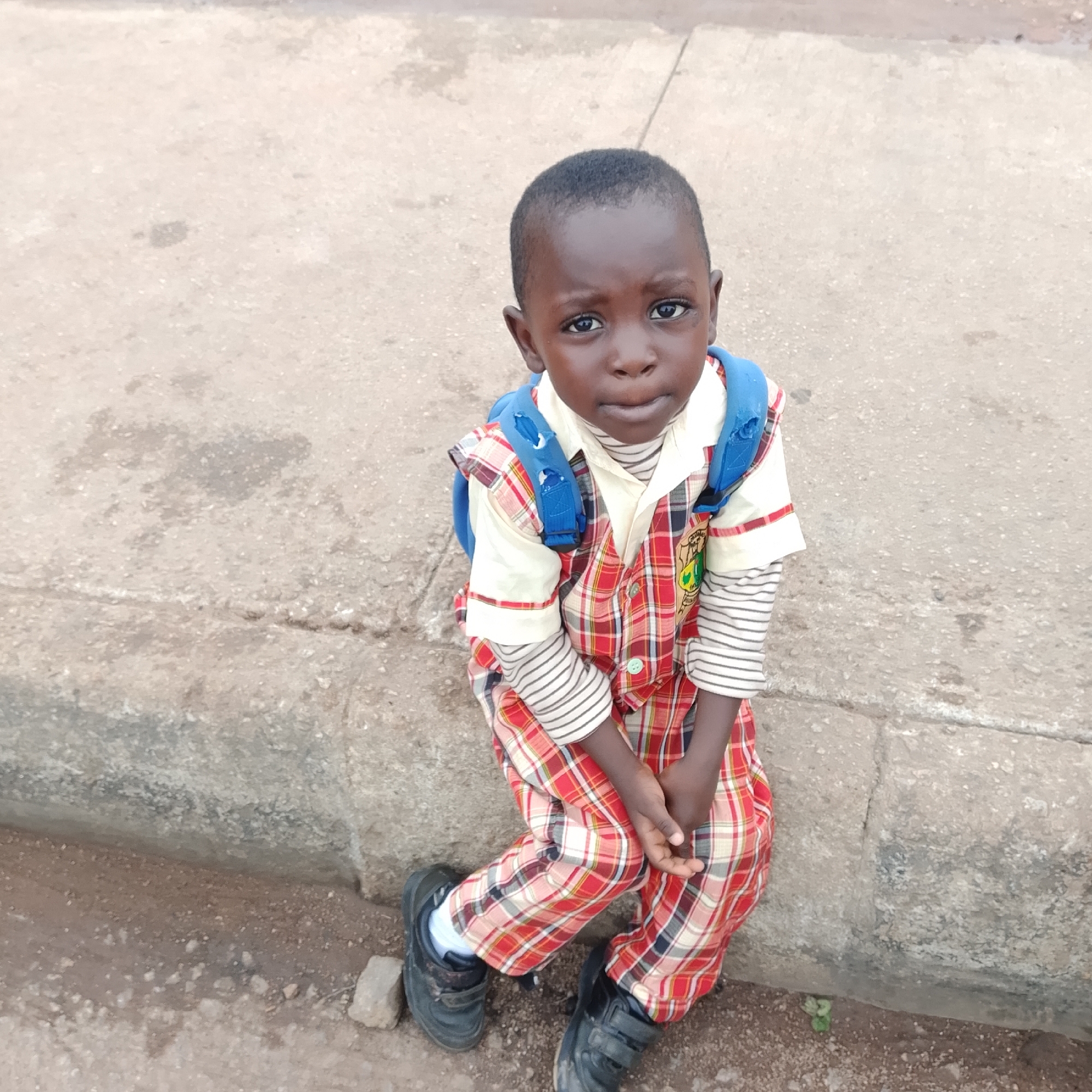 A young boy is sitting on a concrete curb in an outdoor urban setting. He is wearing a red and white plaid outfit over a striped long-sleeved shirt, and a blue backpack. His shoes are black and appear to be velcro closures. The ground is a mix of concrete and dirt. The image was taken in Gura Top, Nigeria.