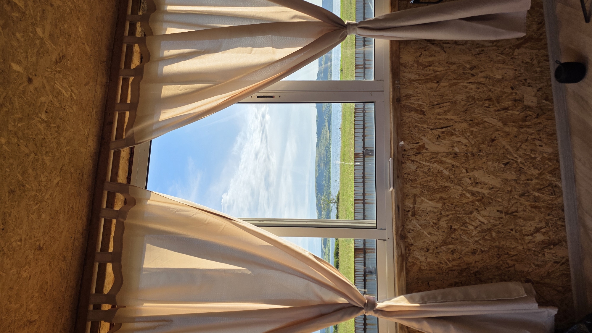 This media file depicts a view from inside a room with walls made of oriented strand board (OSB). A large window, framed by beige curtains tied back with fabric bands, offers a view of a bright, sunny day. The sky is a vibrant blue with scattered white clouds. Beyond the window, a landscape unfolds featuring a body of water, possibly a lake or river, bordered by rolling hills covered in green vegetation. A dark fence runs along the edge of the grassy area in front of the water. The overall scene suggests a rural or natural setting.
The OSB walls and simple curtains imply a rustic or unfinished interior, possibly a cabin, shed, or a room under construction. The bright daylight indicates it is daytime, likely late morning or early afternoon, given the quality of light. The weather appears to be clear and pleasant. There are no people visible in the image. The scene evokes a sense of peace and tranquility, looking out onto a beautiful natural vista. The location is identified as Kupoyarovo, Russia.