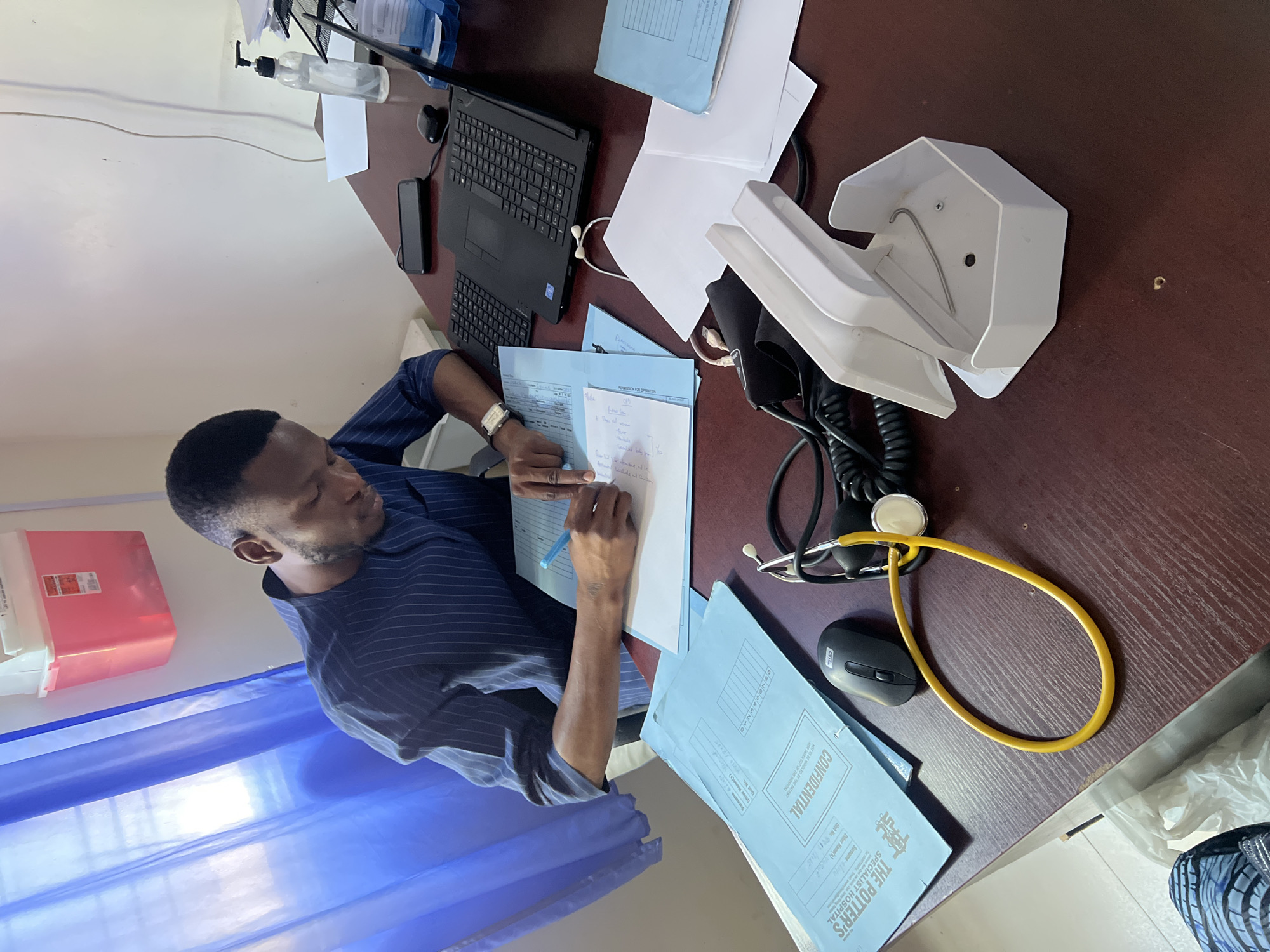 A man sits at a desk in an office. The man is dark-skinned, with short hair and a beard, and is wearing a striped shirt. He is writing on a document with a blue pen. On the desk is a laptop, a phone, a telephone, a computer mouse, a stethoscope, a bottle of hand sanitizer, paper, and blue folders with the words "CONFIDENTIAL" and "THE POTTER'S SPECIALIST HOSPITAL" printed on them. A sharps container hangs on the wall behind him. A blue curtain is partially visible. There is no explicit evidence to confirm the city of Jos, Nigeria, as the location.