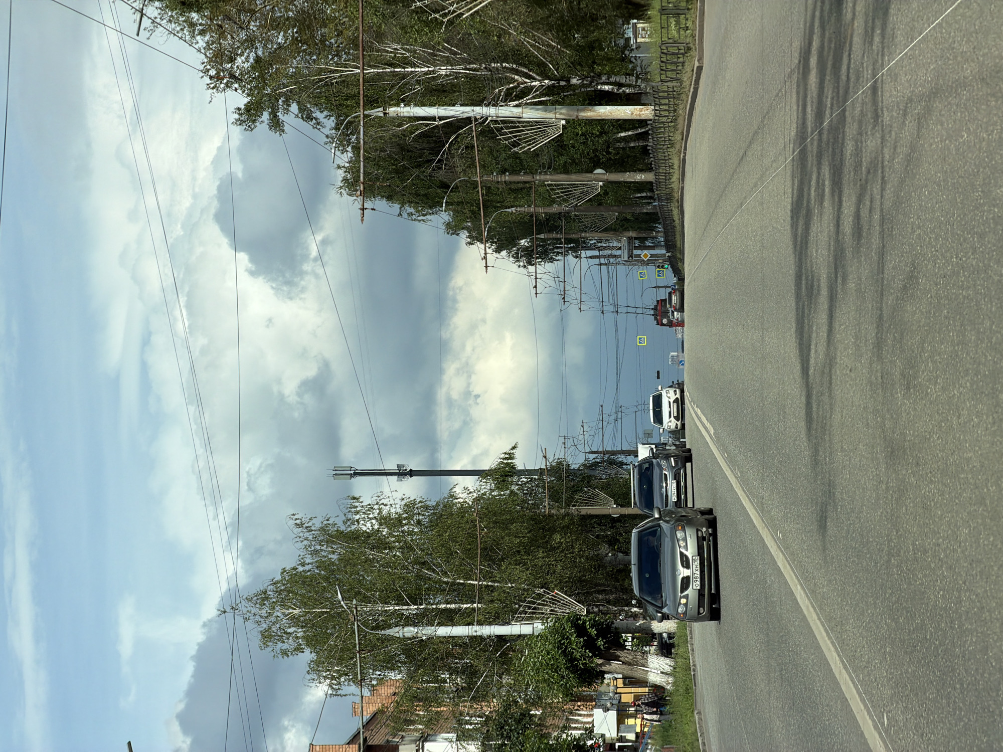 The image shows a roadside view, likely in a town or city. The perspective is from inside a vehicle traveling on a paved road. The road is lined with trees and utility poles supporting numerous wires. In the distance, there are buildings visible.
The main subjects are the cars on the road. A silver sedan is in the foreground, followed by a dark SUV, and then a white car further down the road. There are no visible people. The scene appears to be during the daytime, with a mix of bright sunshine and cloudy skies, suggesting a partly cloudy or overcast day.
Notable details include the many overhead wires, indicating an area with electricity and possibly telecommunications infrastructure. The trees are leafy, suggesting it's a warmer season like spring or summer. There are also traffic signs visible along the side of the road. The license plate on the silver sedan is partially visible and appears to read "G087 XU 18". Another license plate on the dark SUV is also partially visible and appears to read "M634MH 77".
The overall impression is a mundane street scene, likely on a regular commute or journey. There are no strong emotional cues conveyed by the image. The setting is a paved road with infrastructure and vegetation, typical of an urban or suburban environment.
