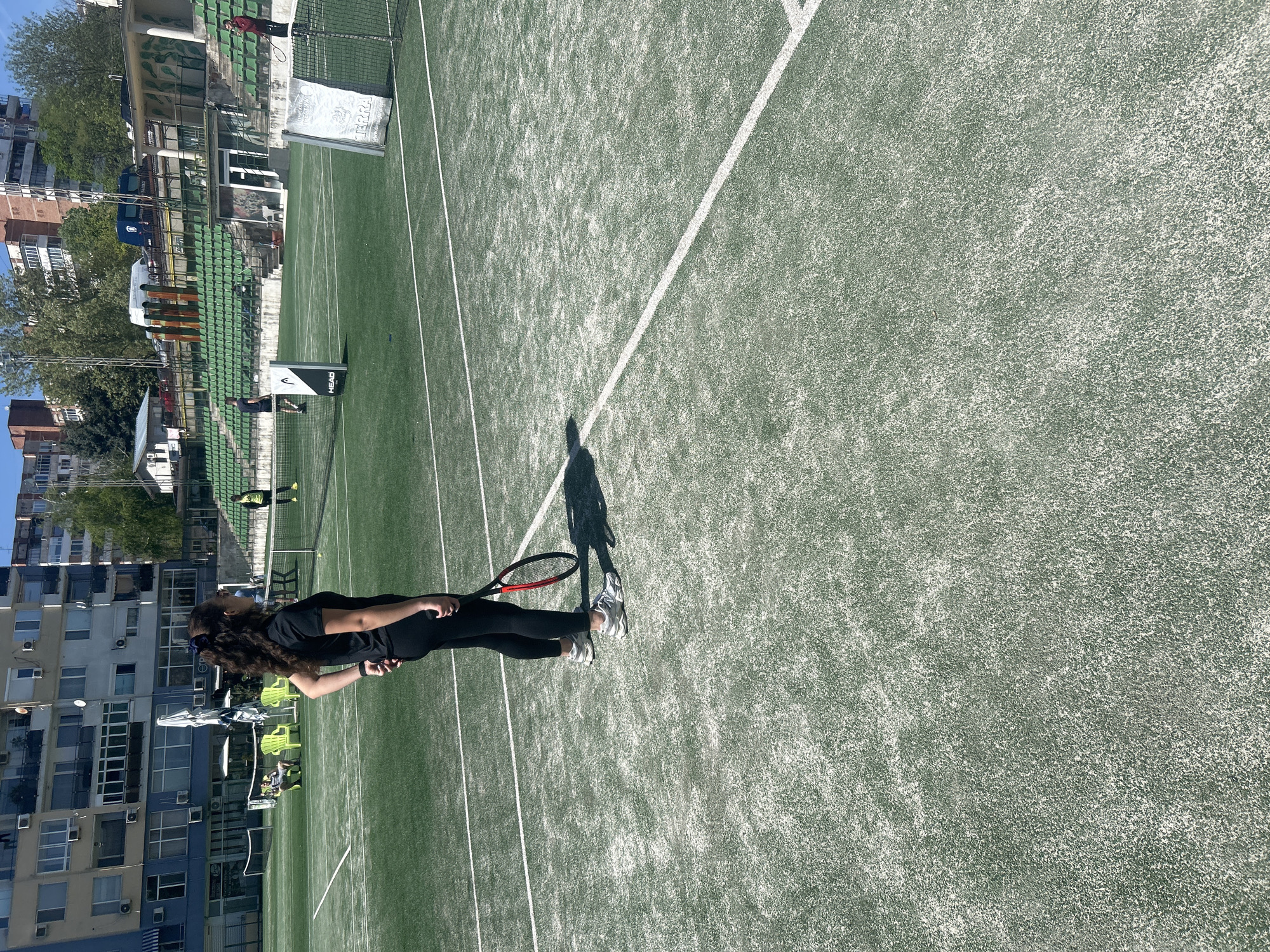 The image depicts a woman standing on a green tennis court. She is wearing dark clothing and holding a tennis racket. The court is marked with white lines. In the background, there are buildings, trees, and spectator seating.
Based on the visible landmarks, the location is in Veliko Tarnovo, Bulgaria. There's an apartment building and a stadium grandstand in the frame. A HEAD sign is visible near the tennis net. The court has an artificial turf.