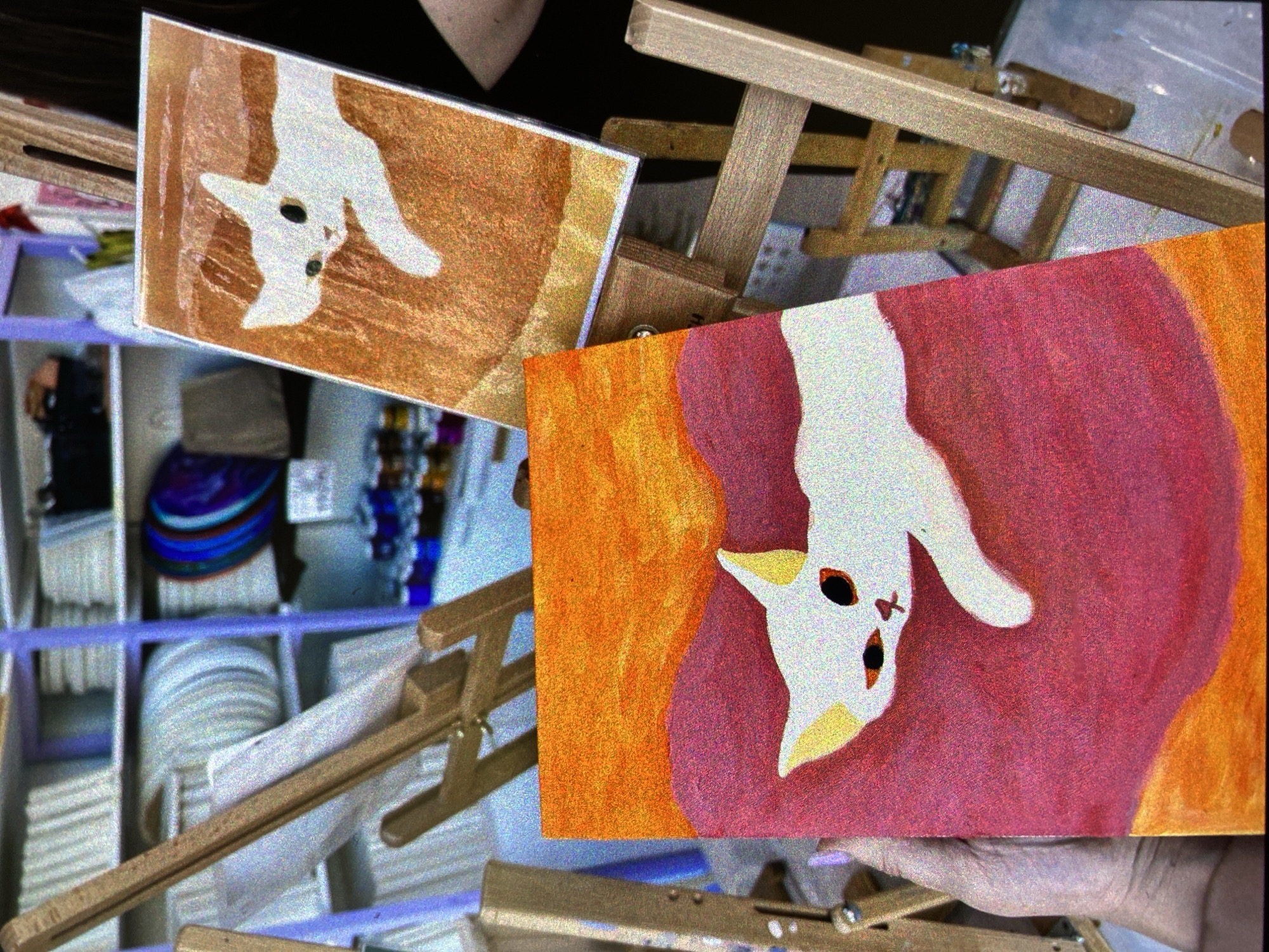 The image shows two small paintings of upside-down cats on easels. One has a primarily brown background, the other, orange and pink. A wooden shelf in the background has assorted objects, including what appears to be stacks of paper plates. The location is likely an art studio or shop in Saint Petersburg, Russia, as suggested by the style of art and the prevalence of similar art scenes in the city. A hand is visible at the bottom right.