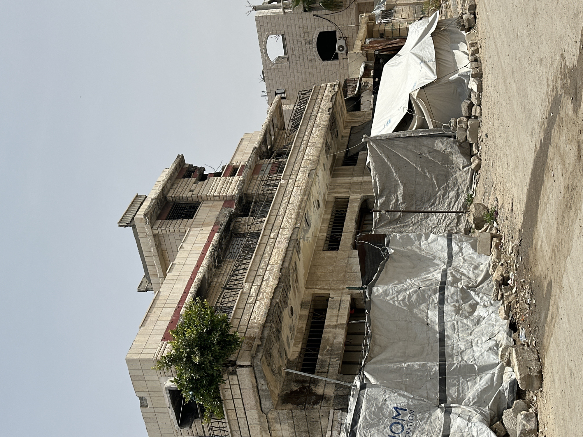 A medium shot depicts a multi-story building in an urban environment. The architecture features pale, potentially stone or tiled construction, exhibiting signs of age and disrepair. The building has multiple windows, some with bars or metal grilles, and balconies with metal railings. There is a small tree growing at the base of the building on the left side.
Adjacent to the building, on the right side of the frame, is a structure covered in what appears to be grey or white tarpaulin. The tarpaulin is secured by rocks and other debris. The ground is bare and rocky, transitioning to a paved surface at the edge of the frame on the right.
The sky is overcast, casting a flat light on the scene. The photograph has been rotated, so the building appears to lean to the left. The image suggests a scene of urban decay, potentially in an area affected by conflict or neglect.