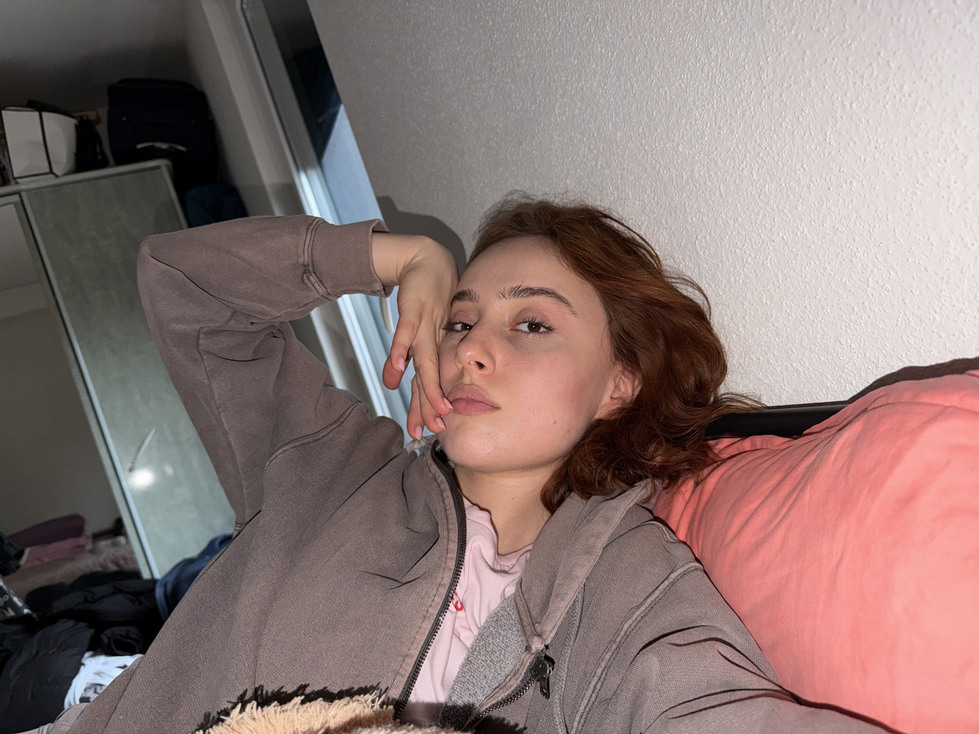 A young person with reddish-brown hair is captured in a close-up, casual pose, lying down in an indoor setting. They are wearing a light pink t-shirt underneath a greyish-brown zip-up hoodie. Their left arm is bent, with the hand resting near their chin and jawline, fingers lightly touching their face. The person looks directly towards the camera with a neutral, perhaps slightly contemplative, expression.
They are leaning against a light peach or pink pillow/cushion on their right side, and a textured, light-colored wall is visible above and behind them. The lighting is artificial and somewhat dim, suggesting an evening or night shot in what appears to be a residential space in Strasbourg, France.
The background on the left reveals parts of a room, including a grey cabinet or wardrobe. On top of this furniture, a white square bag and a dark backpack are visible. Other dark, indistinct items are piled further back, contributing to a lived-in atmosphere. The overall scene portrays an intimate and unposed moment. No visible text is present in the image.