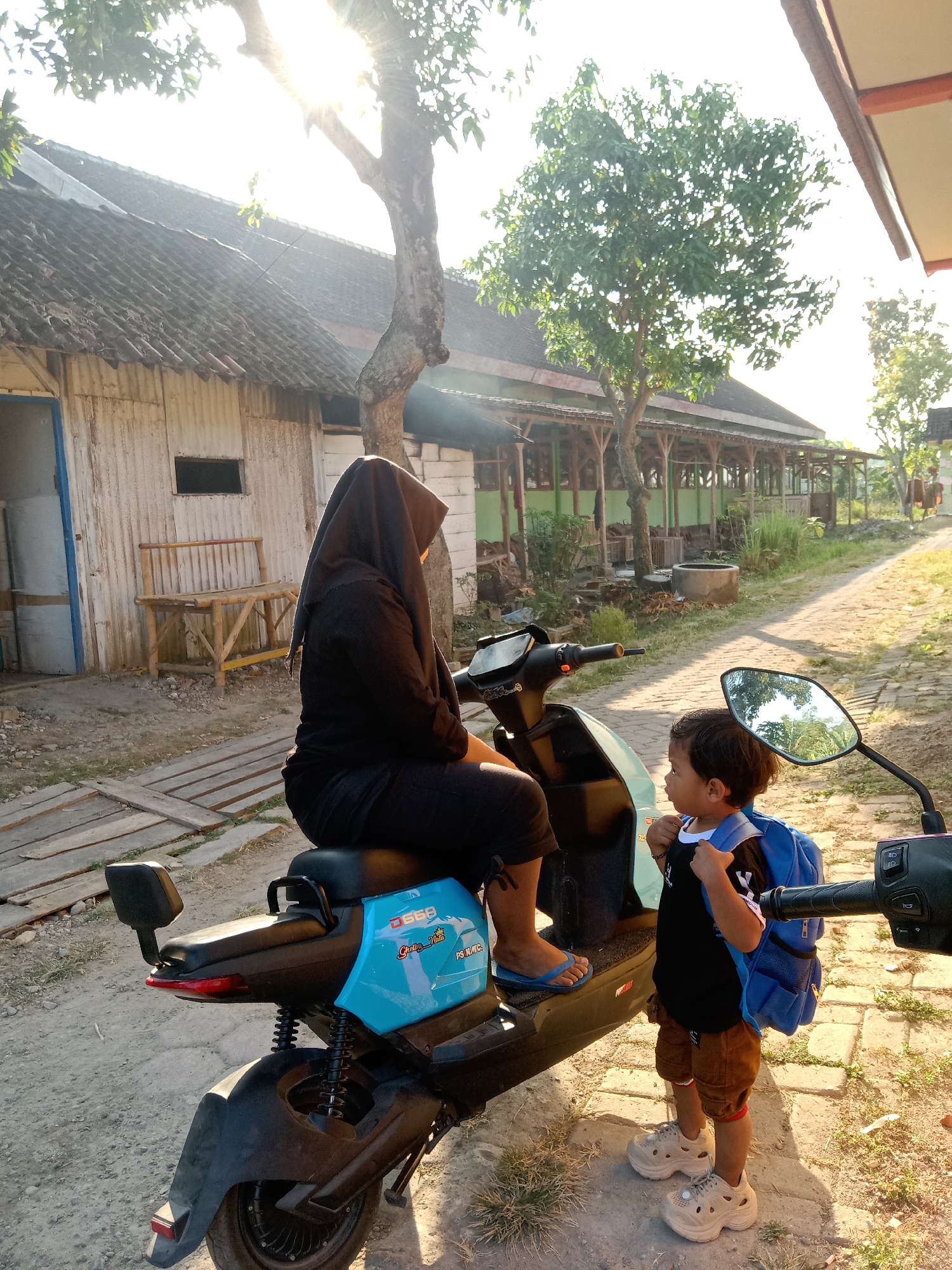 A woman wearing a black hijab and black clothing is sitting on a blue and black electric scooter. A young child wearing a black t-shirt, brown shorts, a blue backpack, and white chunky shoes stands next to the scooter, looking towards the right. The scooter has the brand "Electra Vita" and the model "D568" visible on its side. They are positioned outdoors on a dirt and brick pathway. In the background, there is a rustic building with a corrugated metal exterior and a tiled roof, along with a wooden bench. Trees and vegetation are also present. The scene appears to be in Sugihwaras, Indonesia, under bright sunlight.
