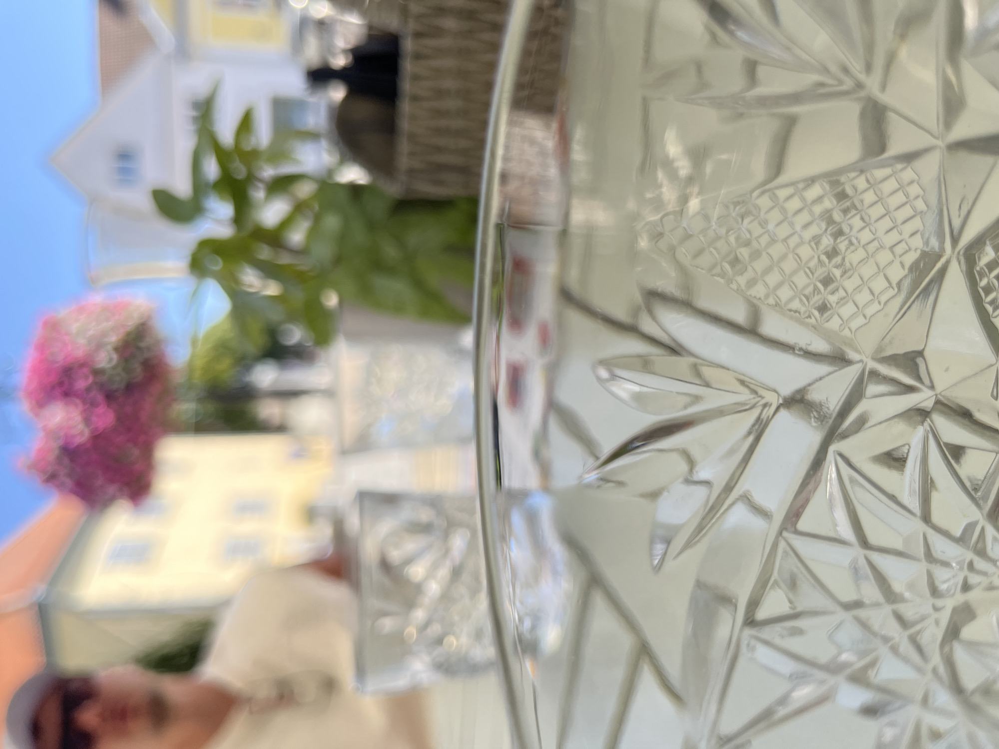An outdoor dining scene is depicted with a blurred foreground featuring a close-up of a clear glass with an intricate etched pattern. Behind the glass, the scene is also out of focus, showing two other glasses, a person wearing sunglasses and a cap, a hanging basket of pink flowers, green foliage, and buildings with orange and white facades. The sky is a clear blue. This scene is consistent with dining at a cafe on a sunny day in Kuressaare, Estonia, where such outdoor seating areas with floral decorations and views of the town's historic architecture are common.