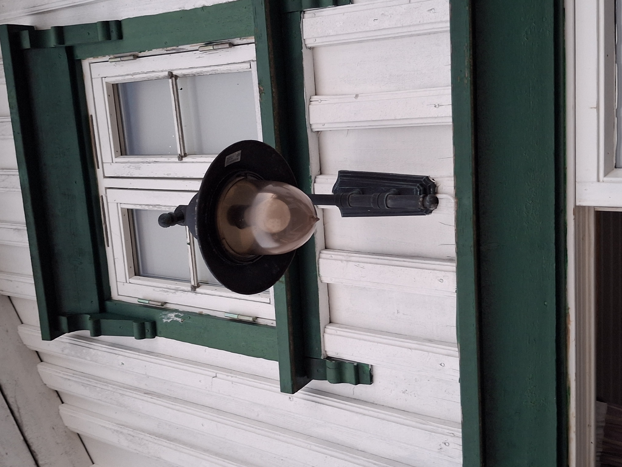 This is a close-up, low-angle shot of the exterior of a white wooden building with decorative dark green trim. A black outdoor lantern with a glass globe is mounted to the wall to the left of a double-paned window. The window is framed by the dark green trim, which also features decorative wooden brackets. The building's siding is white clapboard, with some visible weathering and wear on the paint. The overall impression is of an older, possibly historic, building in a coastal or nautical setting. This image was taken in Åsgårdstrand, Norway.