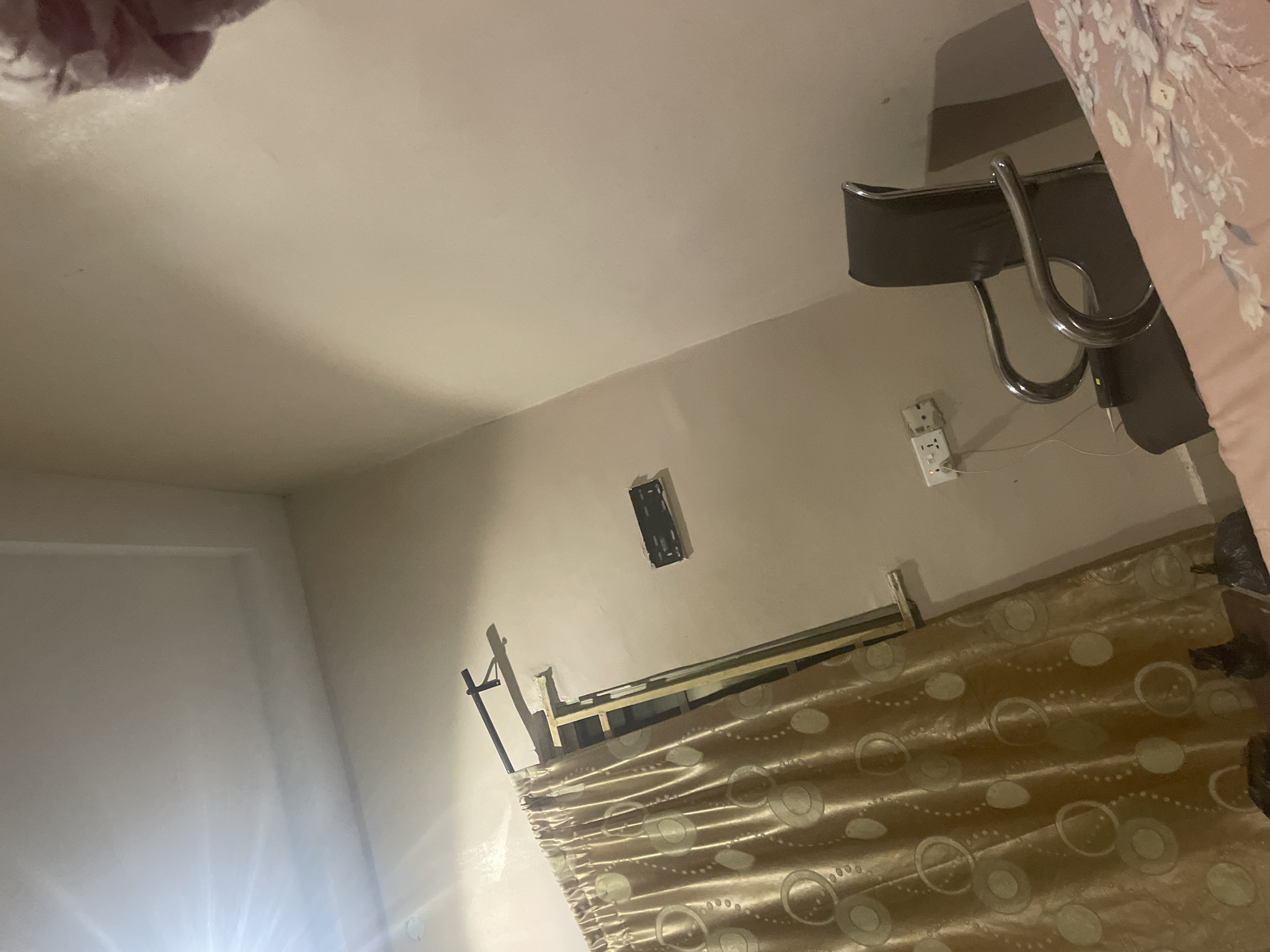 The image depicts an indoor scene, likely a bedroom, in Jalingo, Nigeria. The room has plain, light-colored walls and a white ceiling with a recessed panel. A large window is covered by a patterned golden curtain, hanging from a black rod. Adjacent to the window, a rectangular black object is mounted on the wall. Further down the wall, there are electrical outlets, one of which has a white plug and cord connected. A dark-colored chair with chrome armrests is positioned in the foreground, angled towards the right side of the frame. A portion of a bed with a pink floral pattern is visible in the lower right corner. The lighting suggests it may be evening or nighttime, with a bright light source emanating from the upper left, casting shadows on the ceiling. There are no people visible in the image.