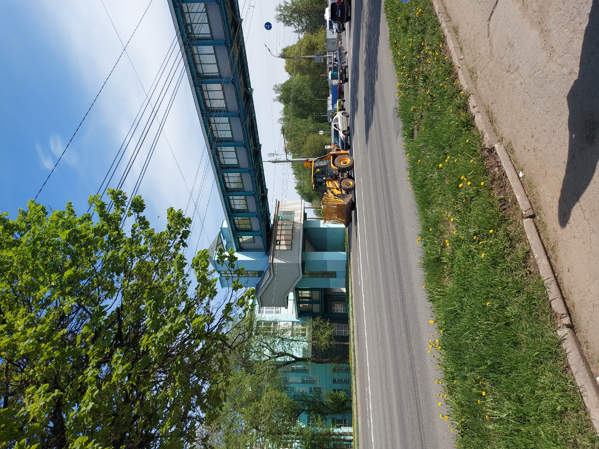 The image depicts a street scene in what appears to be a residential or urban area. A yellow front-end loader is parked on the side of a road. The road is flanked by a grass strip with yellow flowers and a pedestrian walkway on the right. A multi-story building in shades of blue, and a pedestrian overpass is on the left side of the road. Cars are parked in the opposite lane. Tall trees with green leaves frame the foreground and background of the scene. Power lines stretch across the sky, partly obscured by tree foliage and the buildings. The image is taken on a clear day with a blue sky. It may be Izhevsk, Russia.
