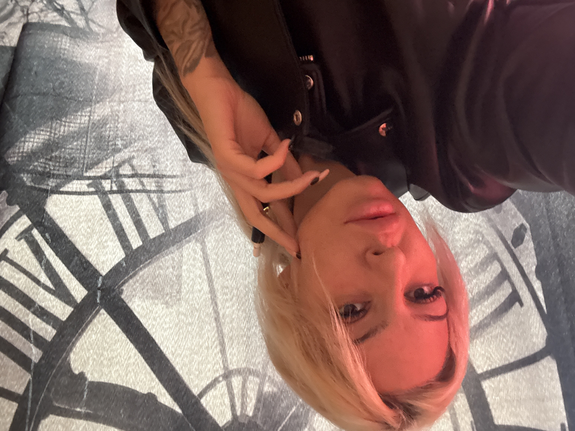 A fair-skinned person with blonde hair, dark eyelashes, and a tattoo on their forearm is looking at the camera. They are wearing a black leather jacket. The person is lying on a surface with a clock design on it. The location is unknown.