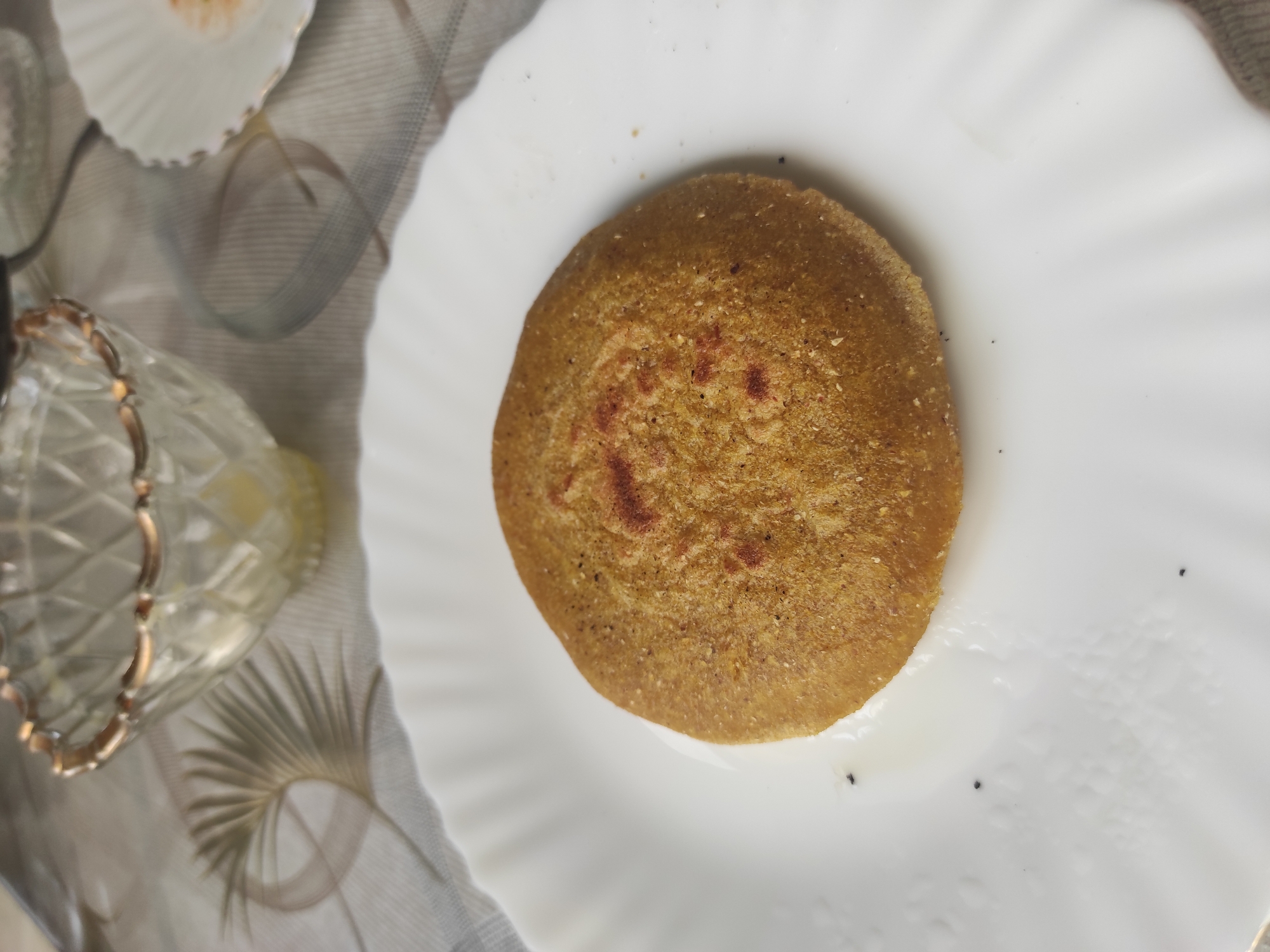 A close-up, overhead shot shows a single, round, golden-brown flatbread resting on a white, fluted plate. The flatbread has a slightly textured surface with some darker, reddish-brown spots. A small amount of liquid, possibly oil or butter, is visible on the plate around the bread. In the background, a decorative, clear glass bowl with a gold rim is partially visible, as is a portion of a white plate with a gold trim, and a patterned tablecloth. The image does not provide any information about the city of Alkhan-Yurt, Russia.