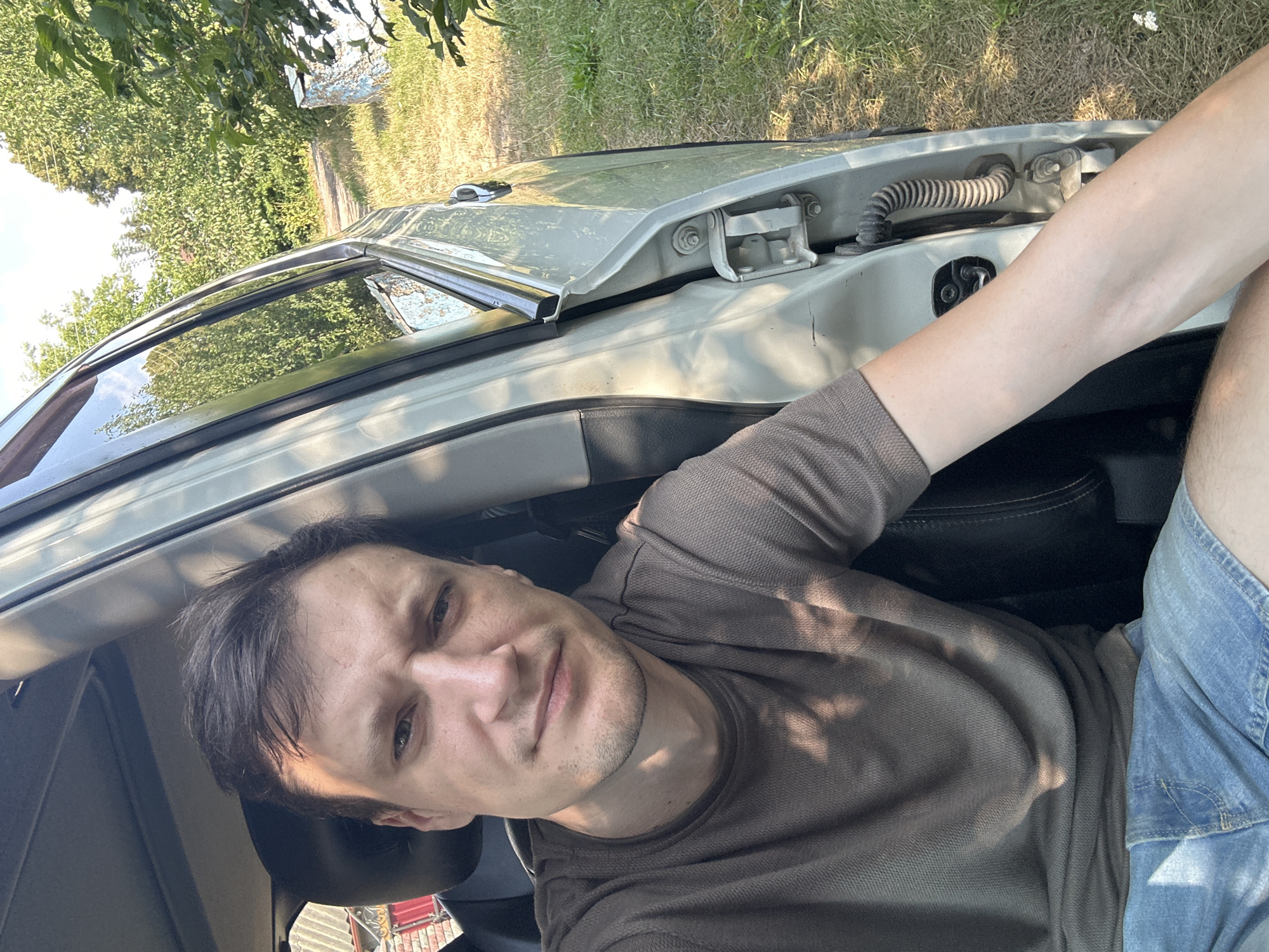 The image shows a man sitting in the driver's seat of a light-colored car, facing the camera. He is wearing a brown t-shirt and denim shorts. The car door is open, revealing the interior and the exterior environment. The exterior consists of a grassy area with trees and a dirt path in the background. The lighting suggests it is daytime, with sunlight casting shadows on the man and the car. The overall scene appears to be in a rural or semi-rural setting, possibly a yard or driveway. There is no specific indication of the city of Shatava, Ukraine within the image itself.