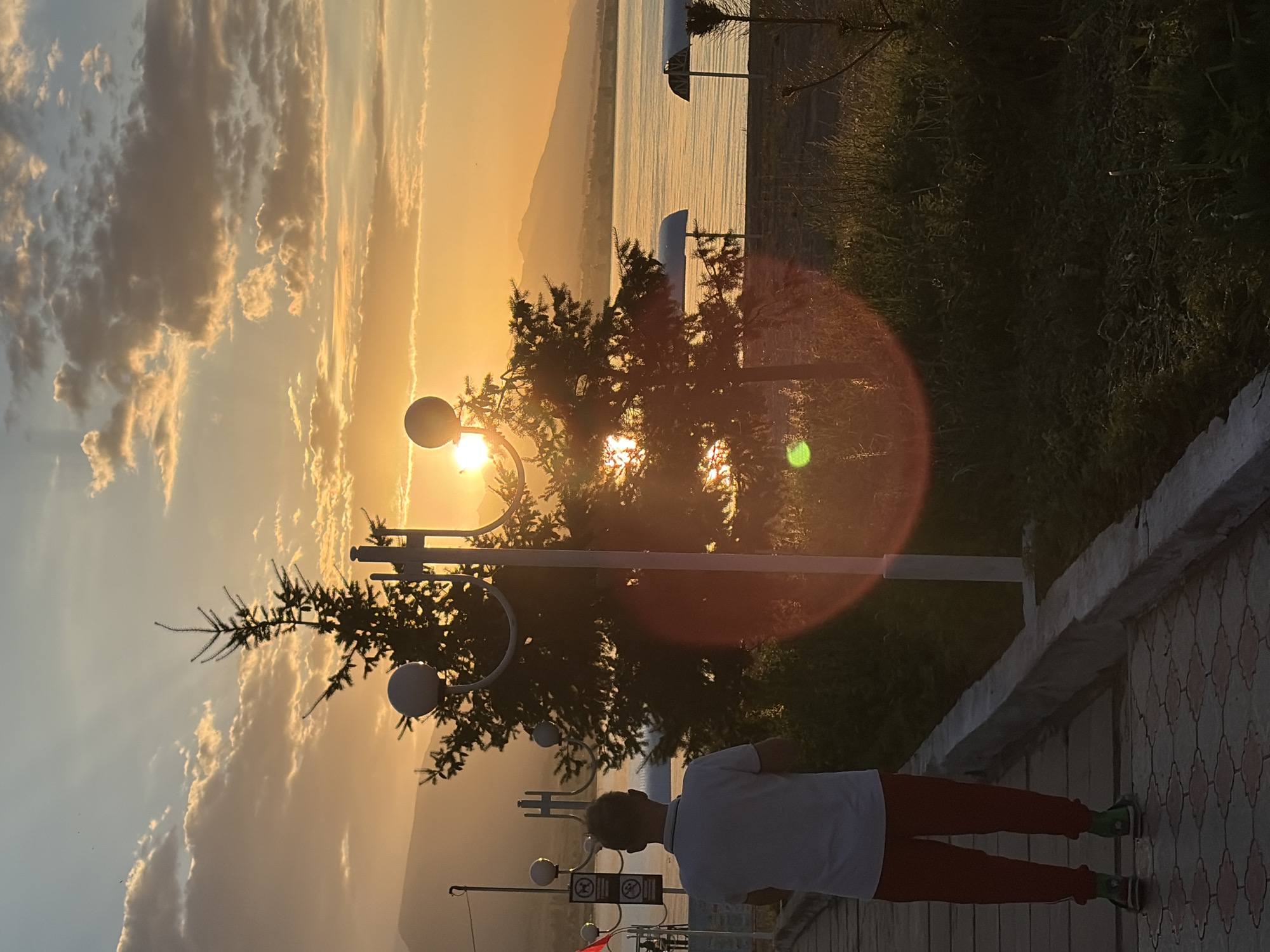 A young person stands on a paved walkway facing away from the camera, looking towards a sunset over a body of water and distant mountains. The person is wearing a white polo shirt and red pants with green sneakers. Two evergreen trees are prominent in the foreground, with lampposts flanking them. The sun is low in the sky, casting long shadows and creating lens flares. The sky is filled with dramatic clouds tinged with orange and yellow hues from the setting sun. The water is calm, reflecting the colors of the sky. Beach shelters are visible on the opposite shore. The ground is a patterned paving stone walkway and a grassy area with scattered foliage.
