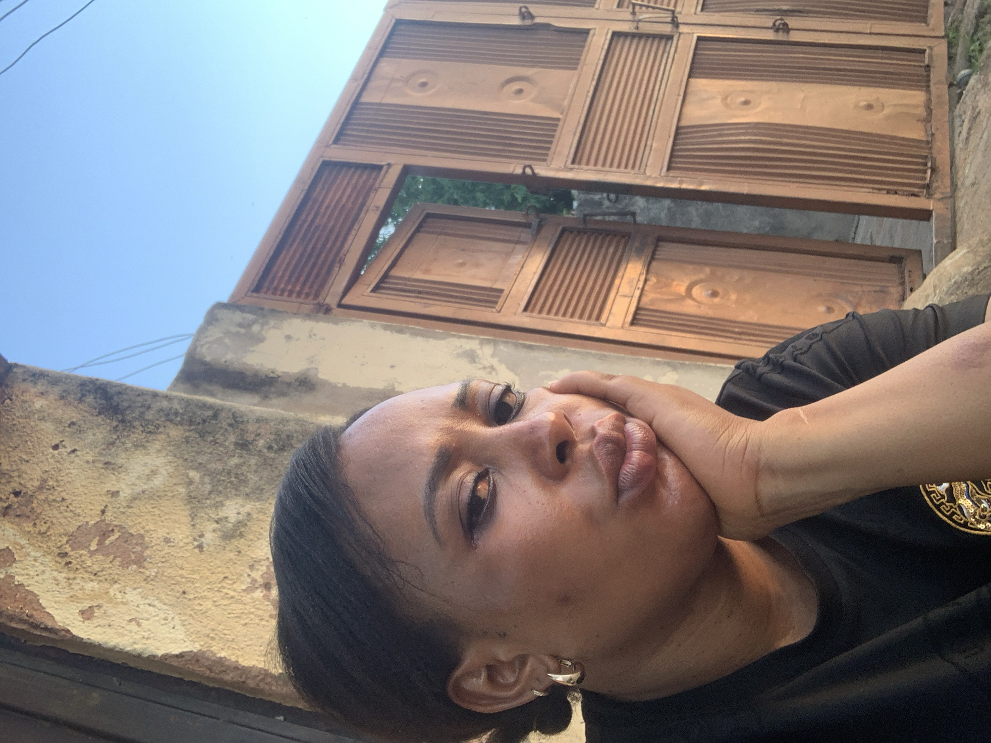 In Sabon Tasha, Nigeria, a woman with dark hair pulled back poses for a selfie outdoors. She is leaning her head on her hand, with her left arm bent and resting on her chest. Her mouth is pursed, and she wears a hoop earring in her left ear. She is dressed in a black t-shirt. Behind her, a weathered concrete wall is visible to the left, and a large, brown metal gate, partially open, stands to the right. The gate is made of slatted panels and decorative circular elements. The sky above is clear and blue. The scene appears to be a residential area.