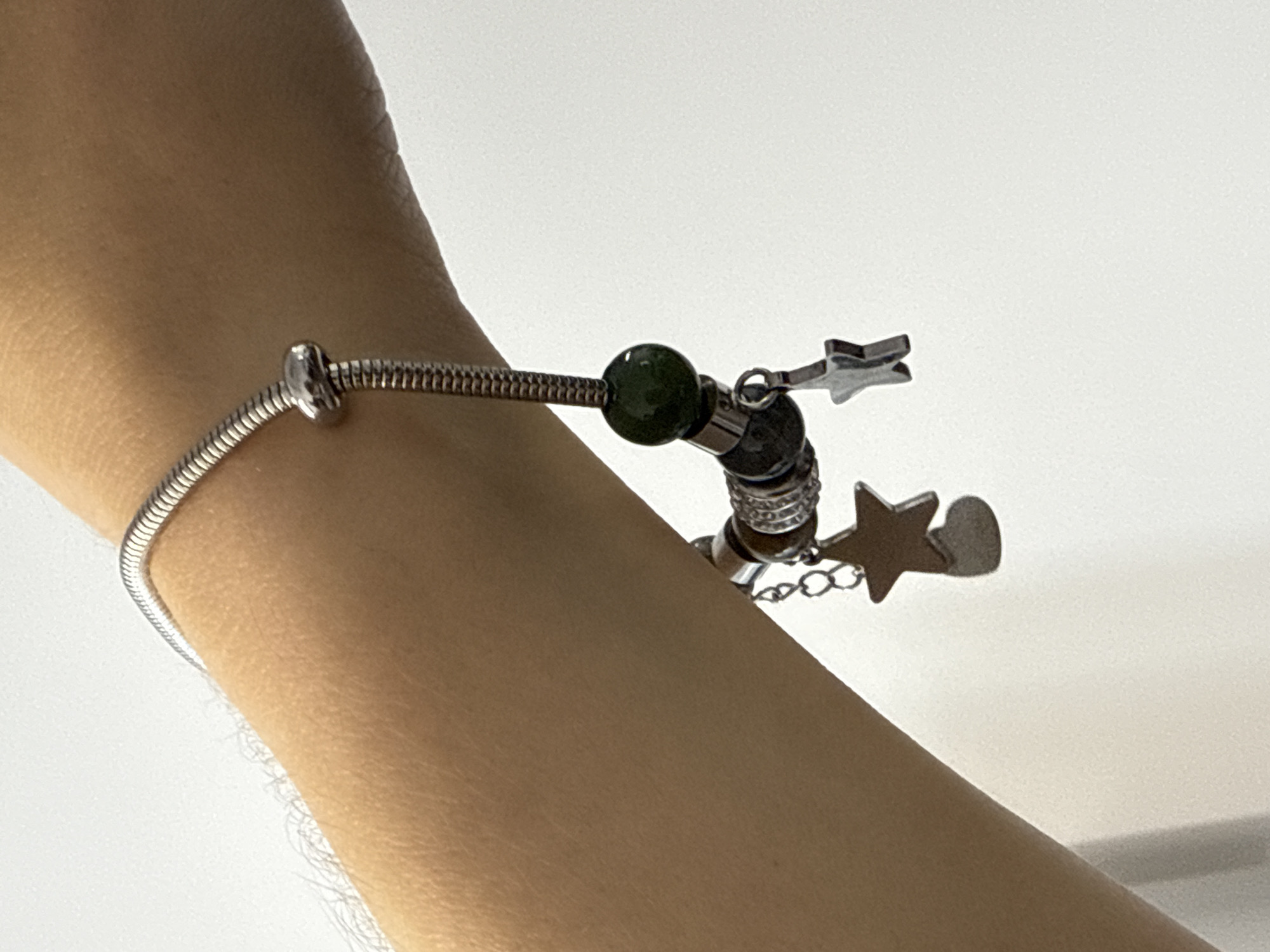 A close-up, eye-level shot captures a person's forearm and wrist adorned with a silver snake chain charm bracelet. The bracelet features a prominent dark green bead, two silver charms shaped like stars, and one silver charm shaped like a heart. The arm displays visible arm hair, and the background is a plain, light-colored wall. The image does not contain information about the city of Łódź, Poland.