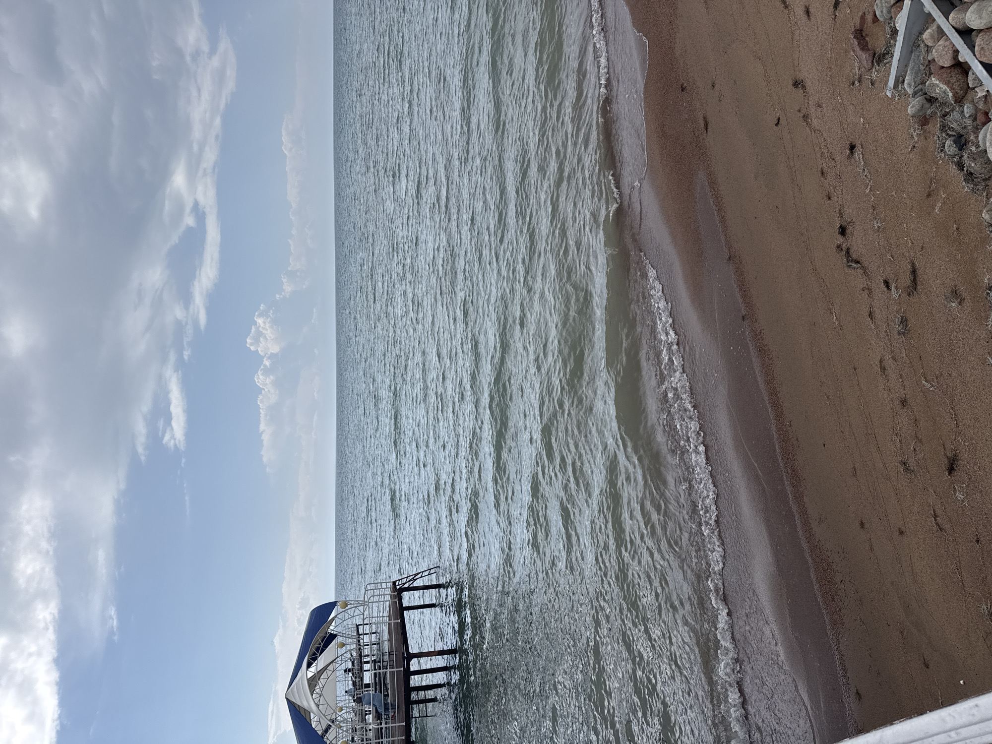 The image shows a view of Issyk-Kul Lake from the shore in Sary-Oy, Kyrgyzstan. A pier extends into the water, with a covered seating area on it. There are a few people visible on the pier, sitting at tables. The water is calm with small waves lapping at the sandy shore. The sky is partly cloudy.