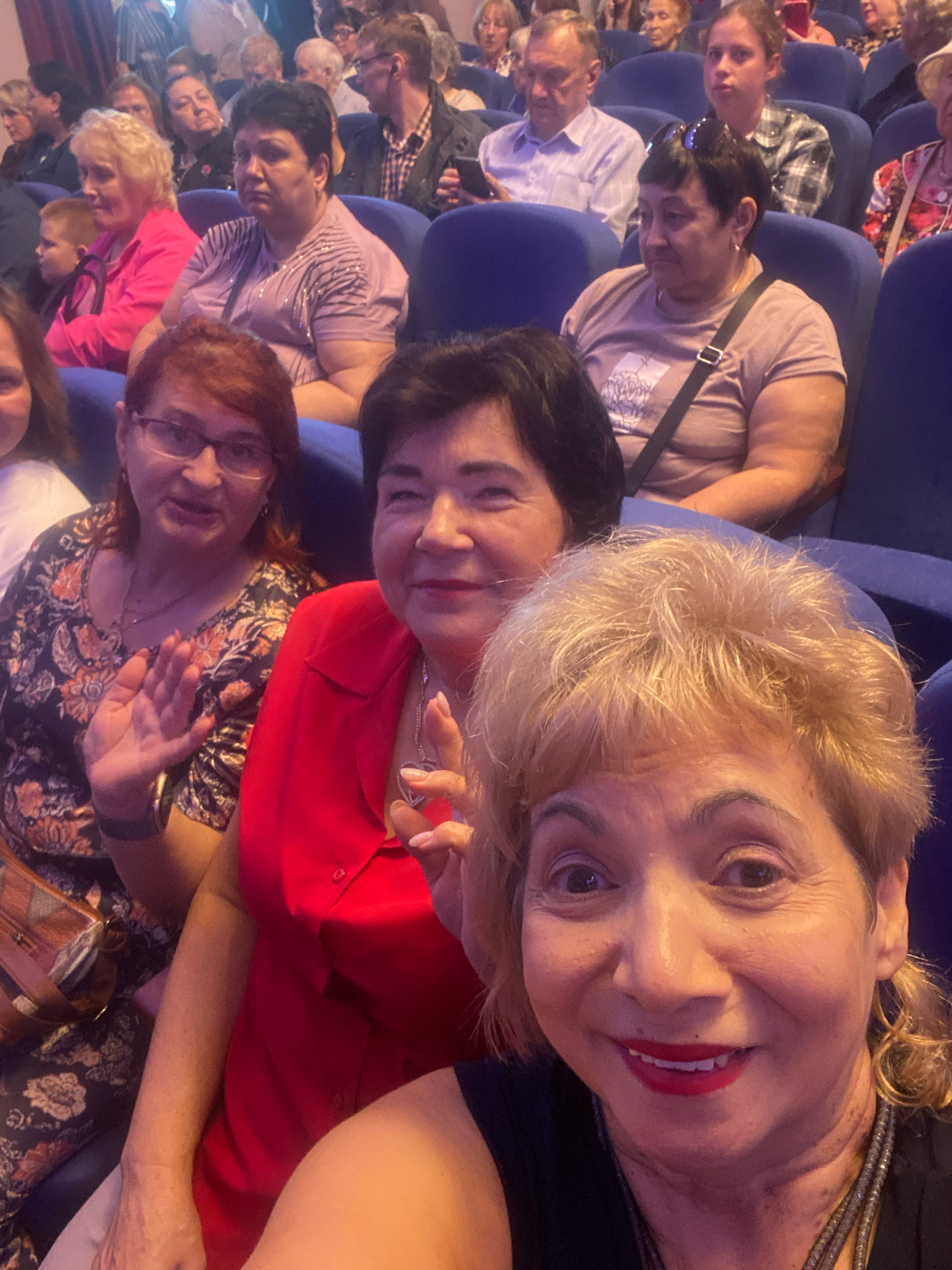 The image shows a group of people sitting in blue seats, likely in a theater or auditorium. The seating arrangement suggests a public event or performance. The foreground features a selfie taken by a woman with blonde hair and red lipstick, smiling broadly. Behind her, other individuals are visible, some looking towards the camera and others engaged in their own activities. The overall atmosphere appears casual and social, possibly during a break or before an event. The location in Ivanteyevka, Russia, suggests a local setting for this gathering.