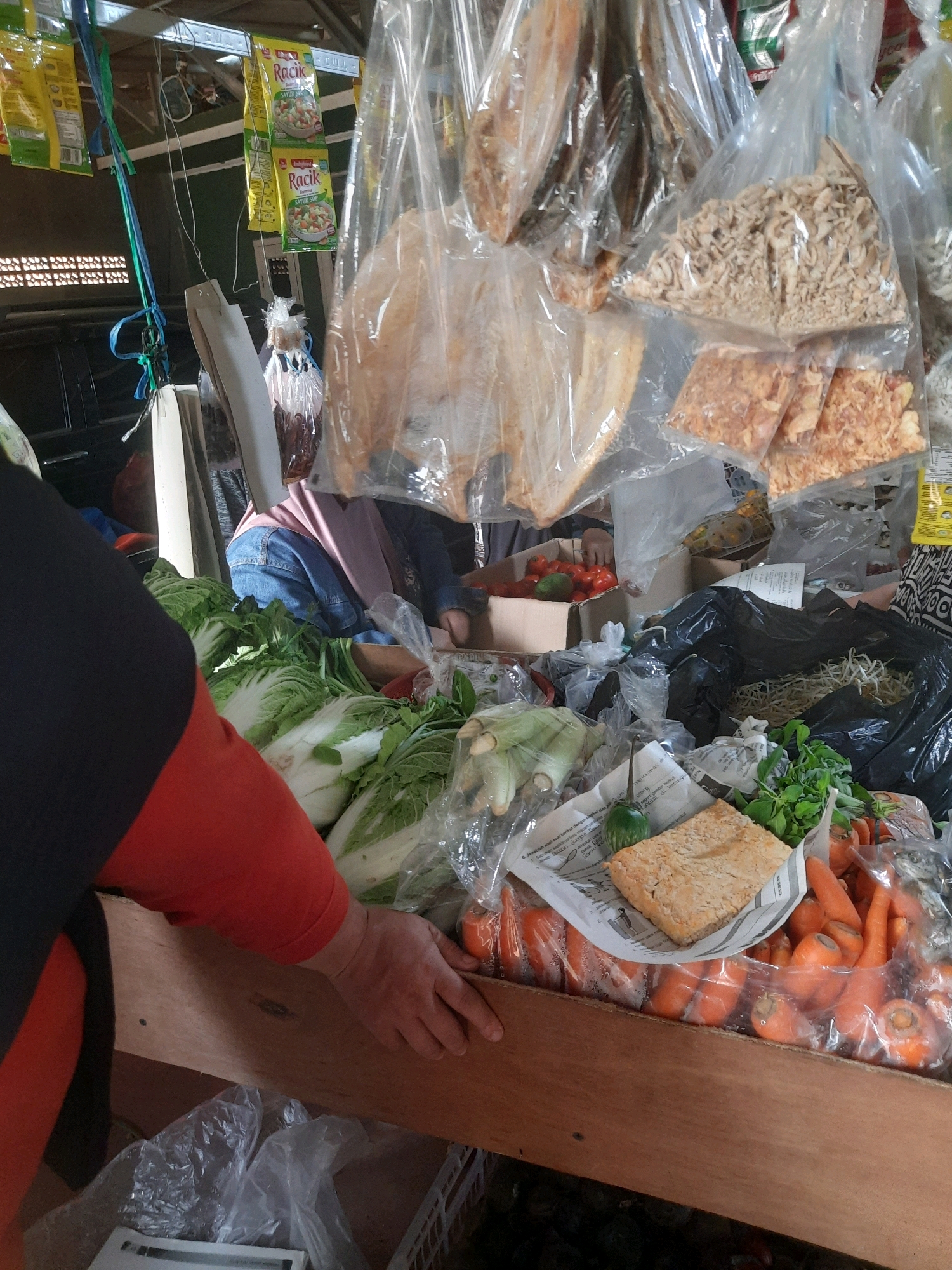 The image depicts a street-side market stall, likely in Gandoang, Indonesia. The stall is laden with various food items, primarily fresh produce and packaged goods. In the foreground, a person wearing a black and orange shirt is reaching towards a display of fresh vegetables, including napa cabbage and carrots, which are packaged in plastic. Behind the vegetables, there are boxes containing tomatoes and other unidentifiable items. Hanging from the ceiling, there are several packaged goods, including what appear to be dried fish and seasoning packets. The overall environment suggests a bustling market setting with a variety of goods for sale.