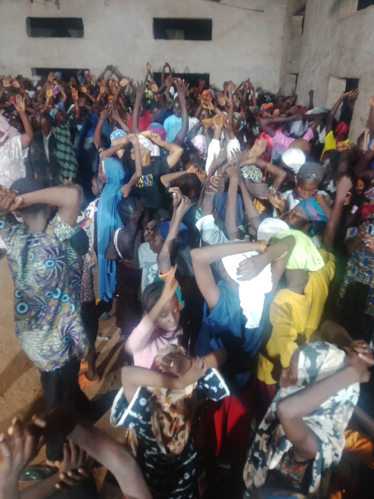 A crowded indoor scene shows a diverse group of people, predominantly women and children, with their arms raised in the air. The lighting is dim, suggesting an evening or indoor setting. The walls appear to be unfinished concrete, and the floor is a dirt or sand-like material. The individuals are wearing a variety of colorful clothing, including patterned fabrics, headscarves, and t-shirts. Some people are looking towards the viewer, while others have their faces obscured or are looking upwards. The atmosphere seems to be one of communal gathering, possibly a religious service, celebration, or protest.