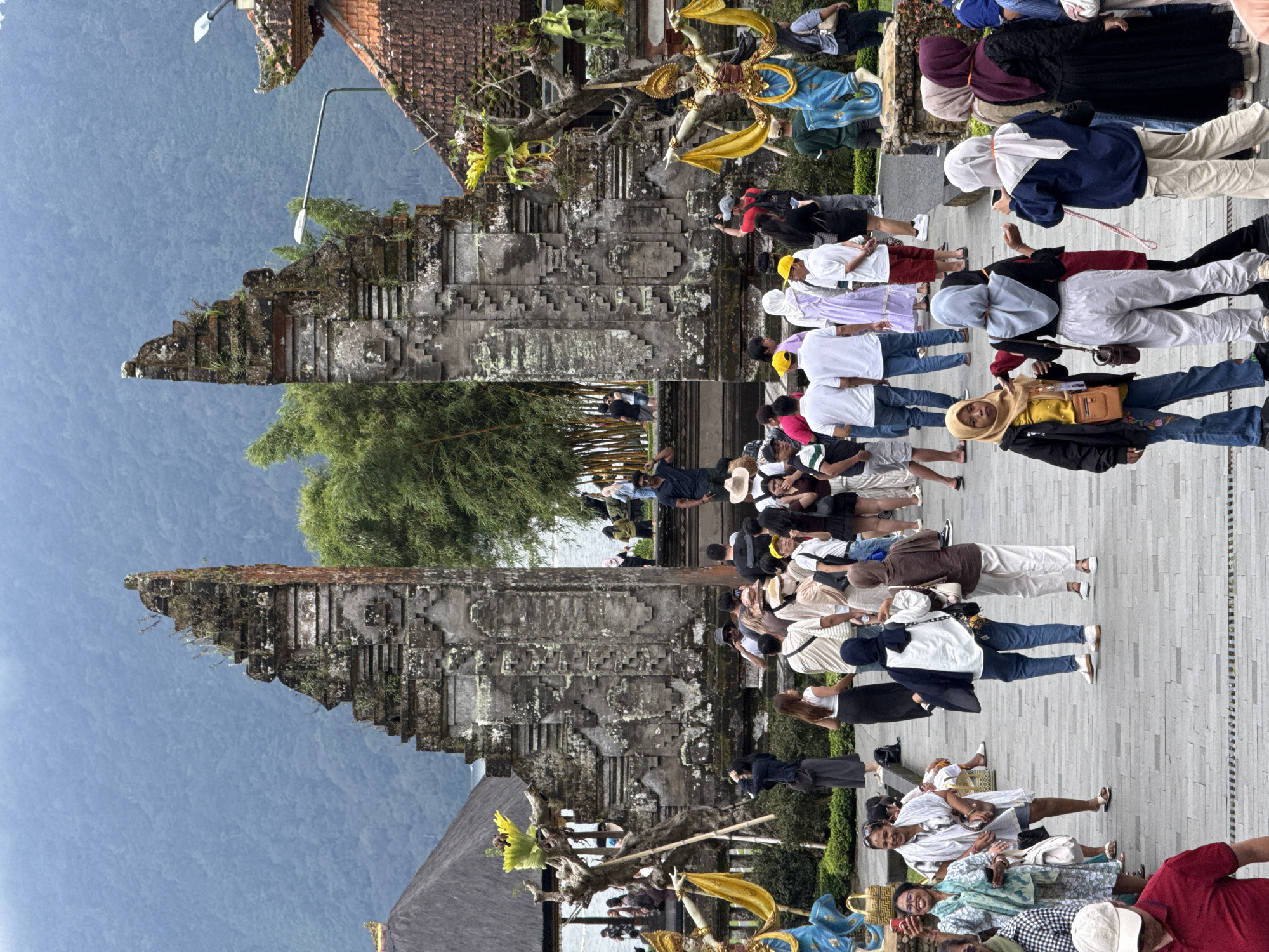 A photograph depicts the Handara Gate in Baturiti, Indonesia, with a group of tourists. The gate consists of two stone towers adorned with intricate carvings and vegetation. A crowd of people is assembled nearby, posing for pictures and interacting with the surroundings. The background features a hazy mountain range, creating a scenic backdrop. The scene appears to be a popular tourist destination.