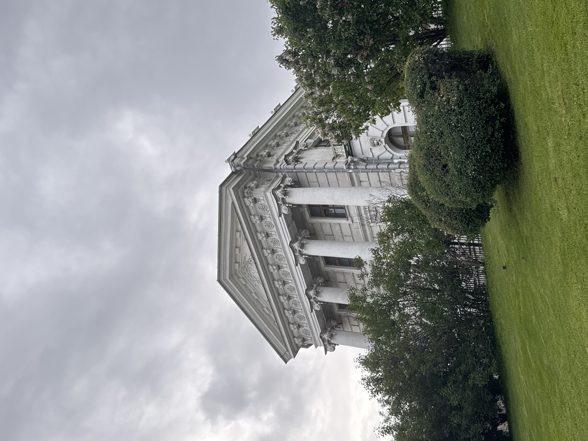 The image shows the Moscow Senate building in the Kremlin, Moscow, Russia. The building is white with neoclassical architecture, featuring columns and a triangular pediment. It is partially obscured by trees and a bush. The sky is overcast, and there is green grass in the foreground.