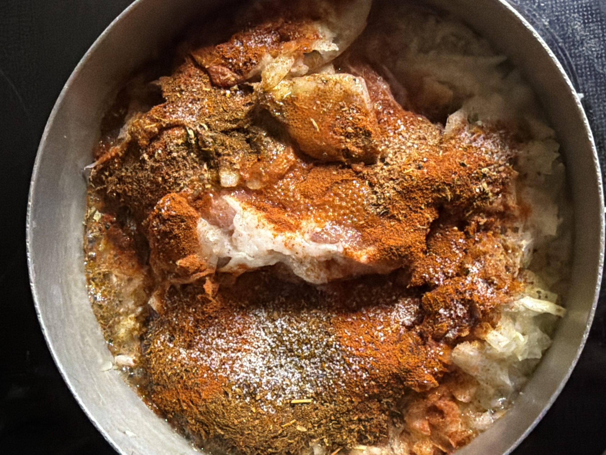 A close-up, overhead shot depicts a metal pot filled with food. The food appears to consist of meat pieces covered in a generous amount of reddish-brown and dark brown spices, possibly including paprika and other dried herbs. Some white, granulated seasoning, likely salt, is also visible sprinkled on top. The meat rests on a bed of what appears to be shredded cabbage or a similar vegetable. The pot sits against a background that transitions from black to a textured, dark grey or blue surface. The location or city is not discernible from the image.