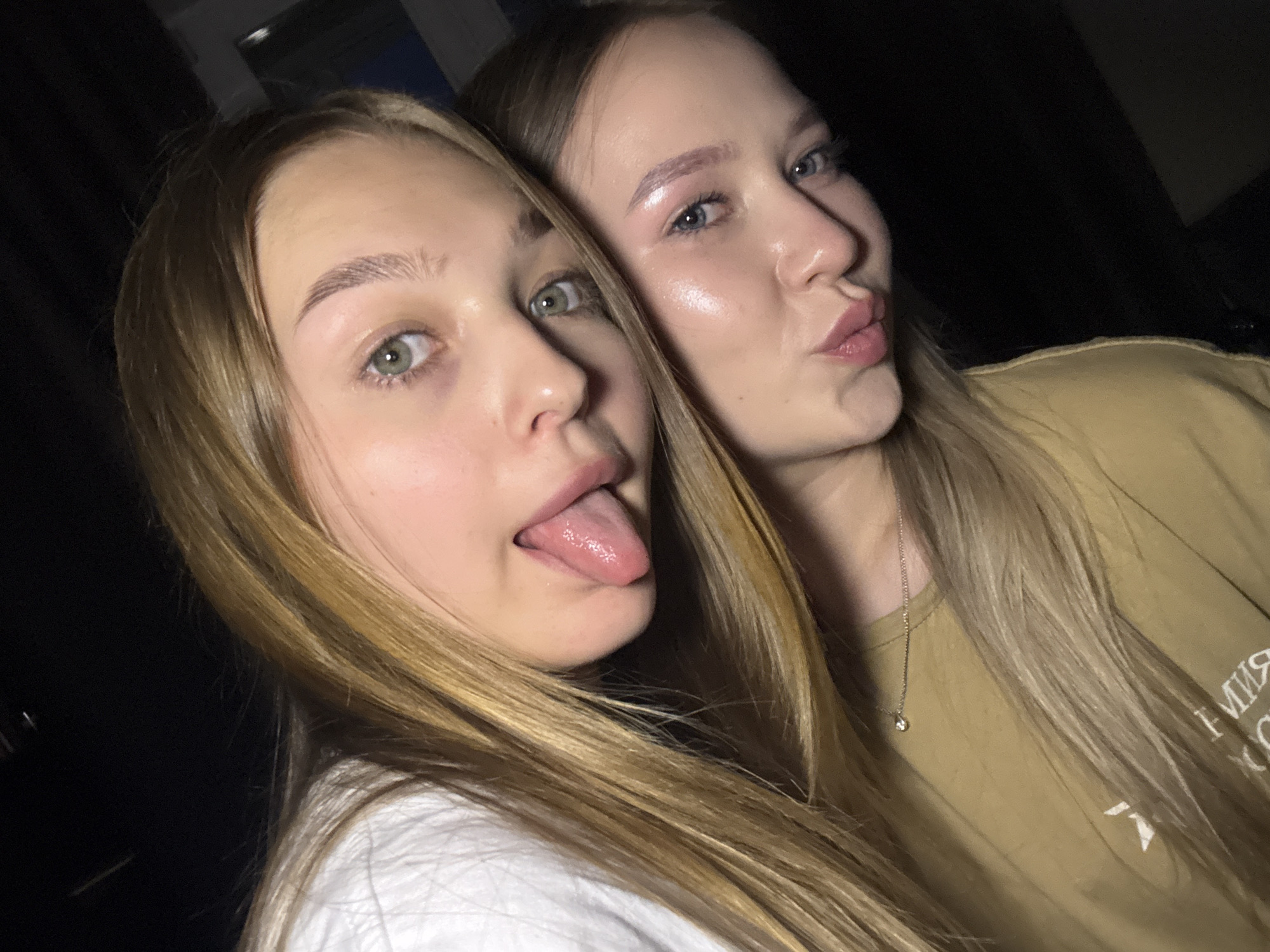 The image is a close-up, selfie-style photograph of two young women, likely teenagers, positioned in a way that their faces are partially overlapping and angled towards the camera. They are both looking directly at the camera. The lighting is dim, suggesting an indoor environment, possibly at night, with the background largely dark and out of focus.
The woman in the foreground, on the right side of the image, has blue eyes and is wearing makeup, including pink eyeshadow and lip gloss. Her lips are pursed. She has blonde hair that falls over her shoulder, and she is wearing a mustard-yellow t-shirt with a faint white graphic and some text in Russian. A thin silver necklace with a small pendant is visible on her neck.
The woman behind her, on the left, has green eyes and is also looking at the camera. Her tongue is sticking out to the side, covered in a slightly glistening substance. Her hair is long and blonde, falling around her face and over the other woman's shoulder. She appears to be wearing a white top.
The image does not contain any identifiable elements that would allow for the specific determination of its location within the city of Chaikovsky, Russia, or any other specific urban area. The scene is primarily focused on the two individuals and their close-up portrait.