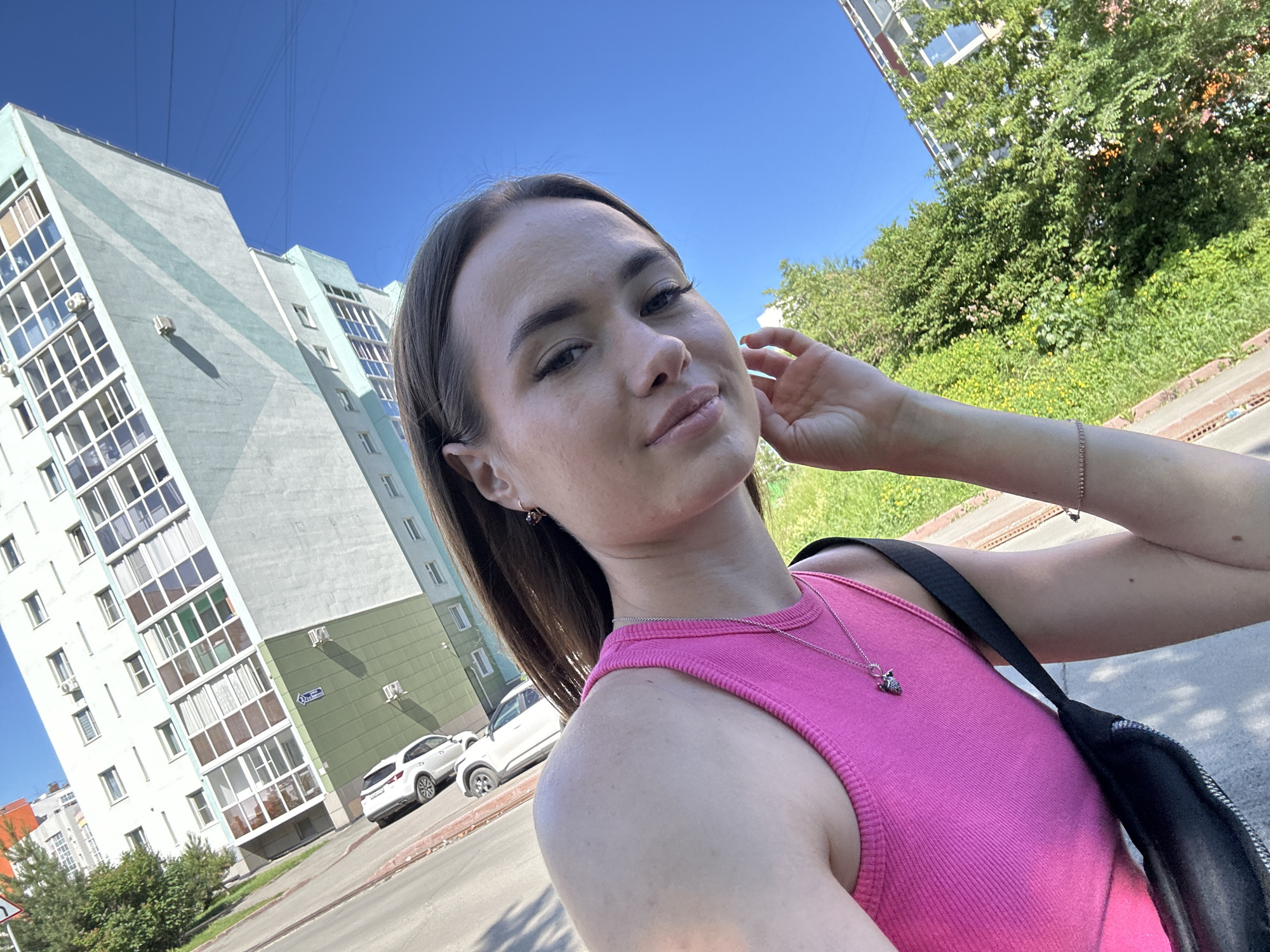 A young woman with brown hair is taking a selfie outdoors on a sunny day in front of a modern apartment building. She is wearing a bright pink sleeveless top and a silver necklace with a small pendant. Her left arm is resting on her head, and she is looking towards the camera with a slight smile. The apartment building is tall and has a light blue and green facade with many windows. A white car is parked in front of the building. The background also shows lush green trees and a clear blue sky. The ground is paved, indicating a street or sidewalk.