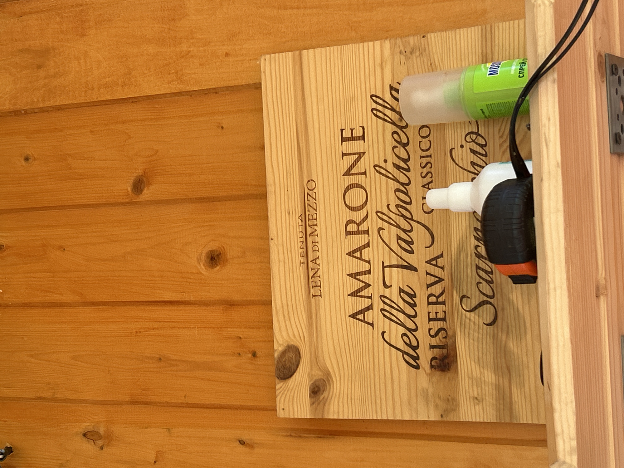 A wooden plank with text carved into it is mounted on a wooden wall. The text reads "TENUTA LENA DI MEZZO AMARONE della Valpolicella RISERVA CLASSICO SCARPACCIO". On a shelf below the plank, there is a white bottle with a spray nozzle, a black and orange object which appears to be a device for blowing air, and a green spray can with Russian text that includes the word "MOSQUITOL" and "СПРЕЙ" (spray). The environment appears to be indoors with wooden paneling. The location is identified as Калицино, Russia.
