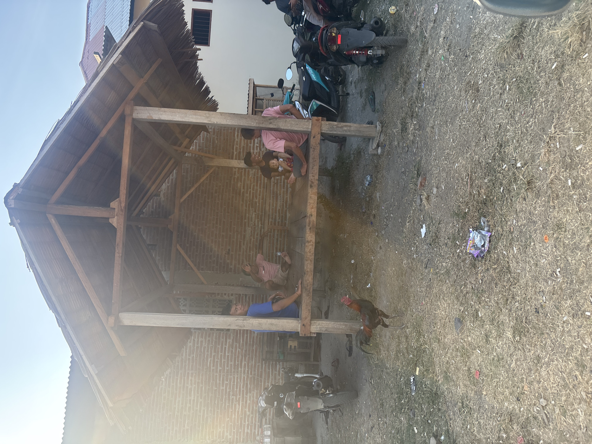 This is a medium-shot, eye-level, outdoor photograph taken in Bunto, Indonesia. The scene depicts a rustic wooden shelter with a thatched roof, which appears to be a gathering place or open-air pavilion. Several individuals are seated on a wooden platform within the shelter. To the left of the shelter, a motorcycle is parked. A rooster is standing in the foreground on the dry, uneven ground, which is littered with debris. The background shows a brick wall and parts of buildings. The lighting suggests it is daytime, possibly late afternoon, with sunlight streaming in from the left, creating lens flare.