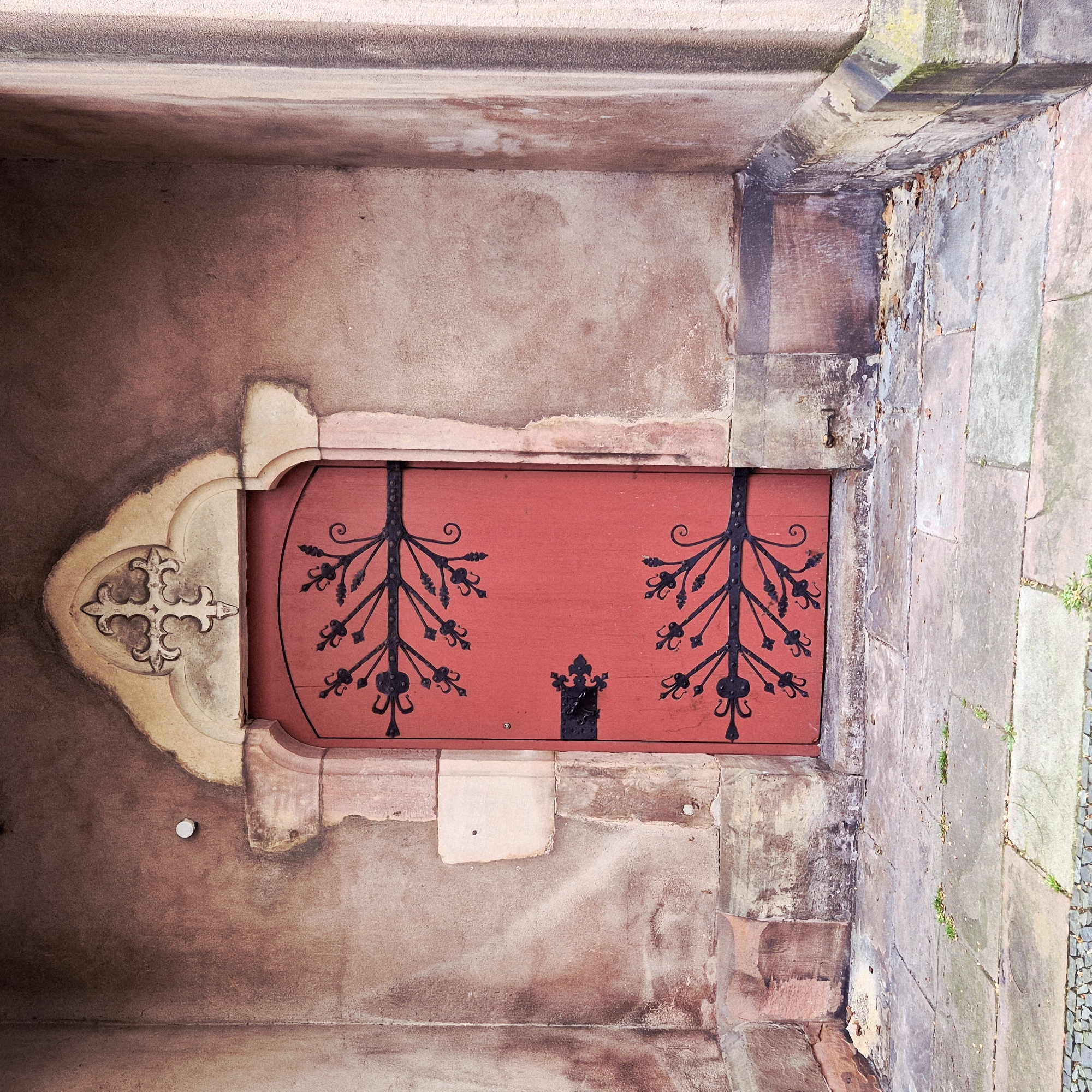 An eye-level, medium shot oriented at an angle reveals an ornate red door, seemingly an exterior door from the city of Strasbourg, France. The door is recessed within a stone or plaster wall. The wall exhibits varied brown hues and textures, suggesting age and weathering. A decorative stone surround with an ornate cross pattern flanks the left side of the door. The red door is embellished with symmetrical black ironwork resembling stylized trees. A simple, black iron latch is located in the center of the door. The surrounding wall features visible stone blocks and patches of plaster, with some sections exhibiting discoloration or wear. A stone walkway or curb can be seen to the right of the door.