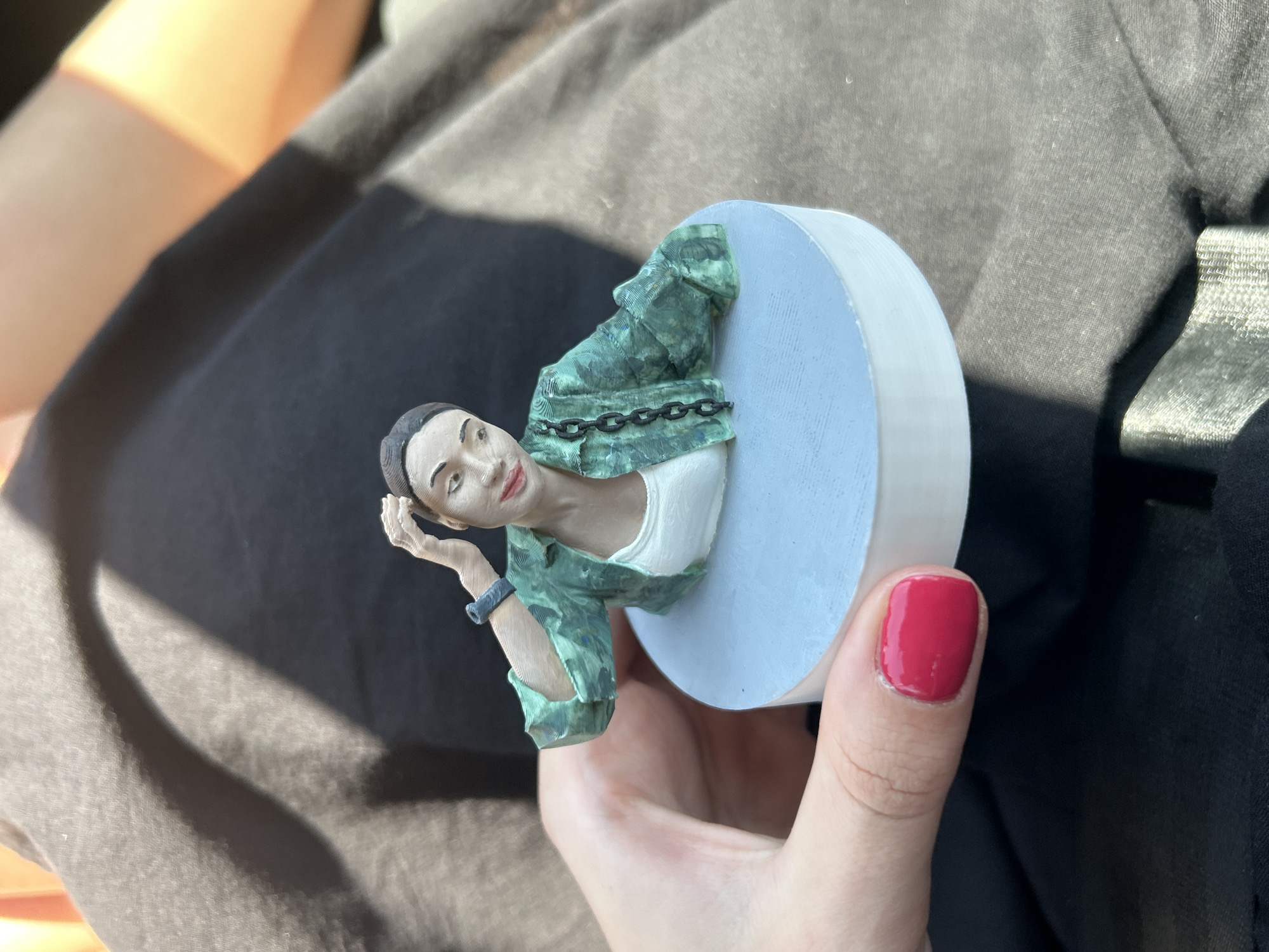 A close-up, slightly elevated shot shows a 3D-printed figurine of a woman with dark hair pulled back, wearing a green patterned jacket over a white top, and a black bracelet on her left wrist. Her right hand is raised to her head, and she is looking to her left with a slight smile. The figurine is mounted on a light blue cylindrical base. A person's hand with bright pink nail polish is holding the base from the left side. The background consists of dark fabric, likely clothing, with dappled sunlight creating shadows and highlights. The image does not provide any information to determine a specific location within the city of Sofia, Bulgaria.