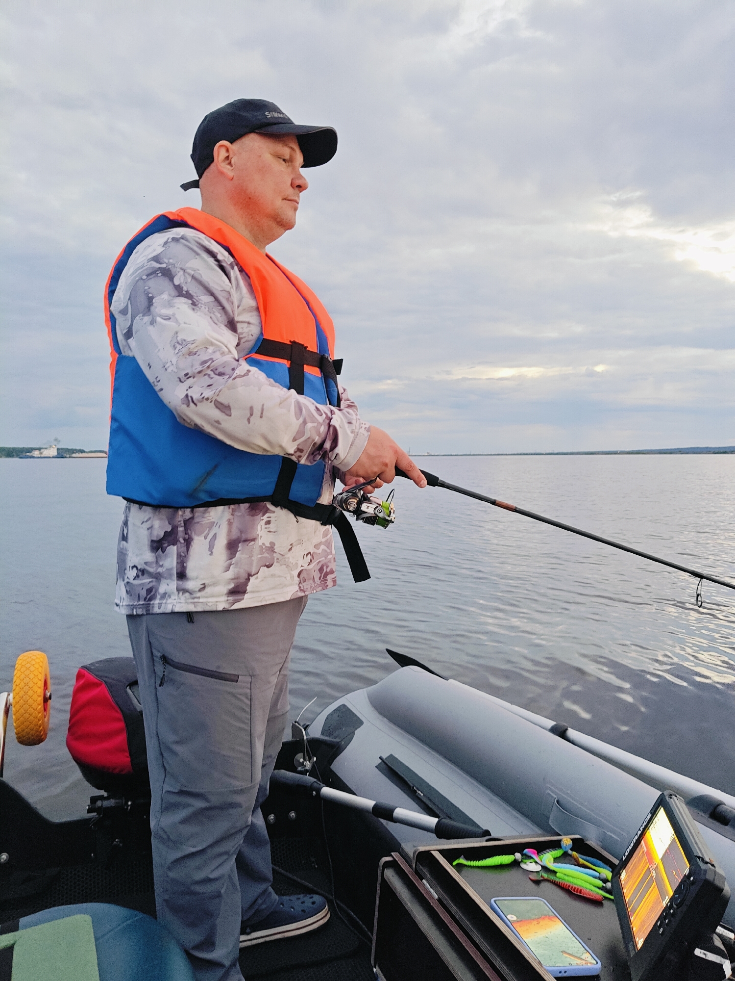 A bald man wearing a black baseball cap, an orange and blue life vest over a camouflage long-sleeved shirt, and gray pants stands on an inflatable boat, fishing. He holds a fishing rod and reel, casting into a body of water. The boat has a red and black bag, a yellow wheel, and various fishing lures and a fish finder on a tray. The background shows a wide expanse of water and a cloudy sky, with a distant shoreline featuring a large building and what appears to be industrial structures, possibly indicating the location is near the Kama River in Chaikovsky, Russia.