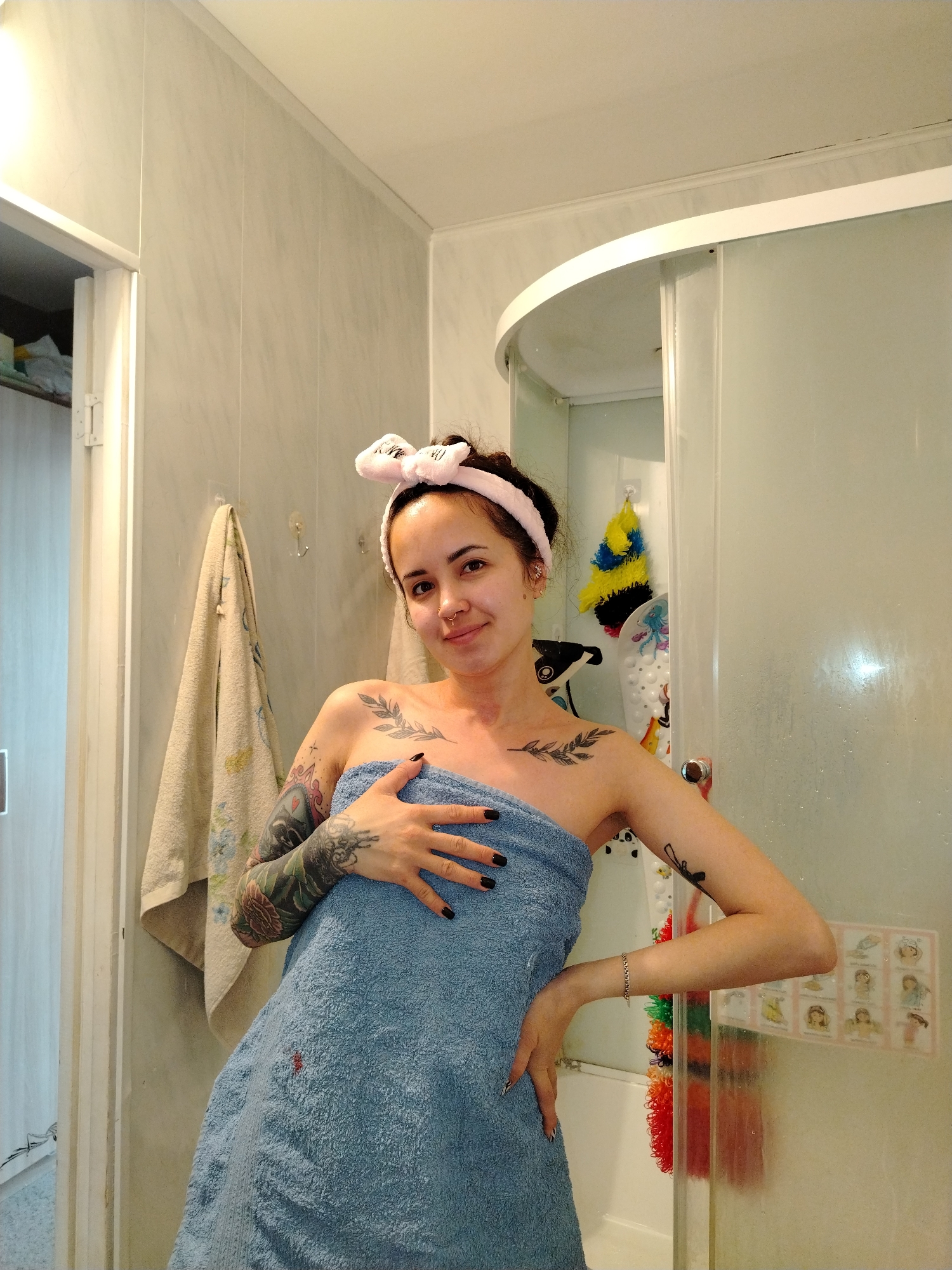 A young woman with dark hair pulled back by a pink headband and tattoos on her arms and chest stands in a bathroom, wrapped in a blue towel. Her right hand is on her hip and her left hand is on her chest. She has dark nail polish and is smiling at the camera. The bathroom has marbled-look paneling on the walls. A towel hangs on the wall to the left. A shower stall with frosted glass doors is behind her to the right. Inside the shower, there is a yellow and blue brush, a white bath mat with a drawing, and a colorful loofah. A white door frame is visible on the left. The bathroom appears to be located in Noviy, Russia, based on possible evidence not immediately clear in the image.
