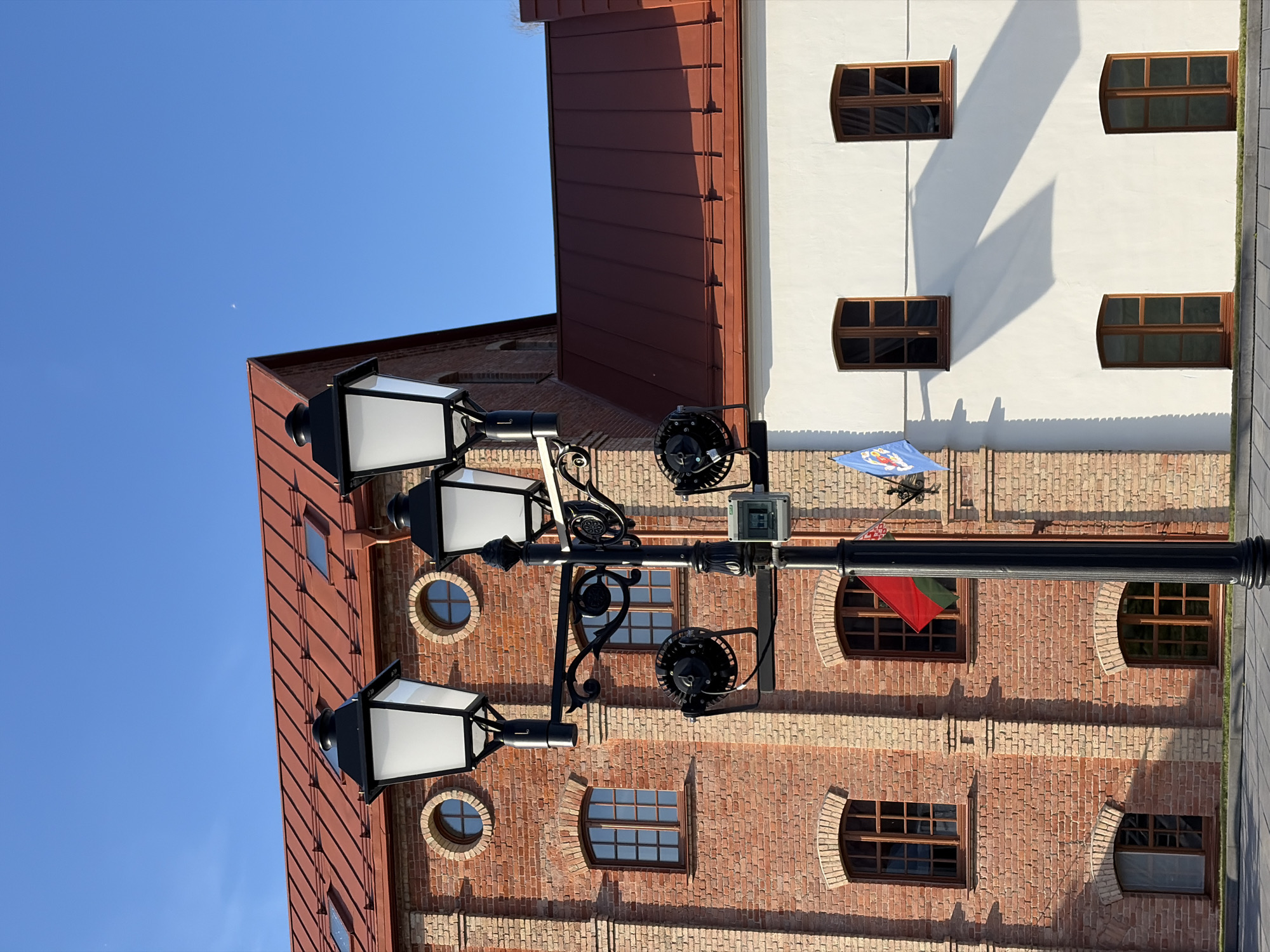 The image shows a street lamp with three lights in front of two buildings with red roofs under a clear blue sky. The building on the left is made of red brick and has several windows and circular windows. The building on the right is white and has several windows. There are two flags on the white building, one red and green and one blue with a crest. The scene is located in the Upper Town in Minsk, Belarus.
