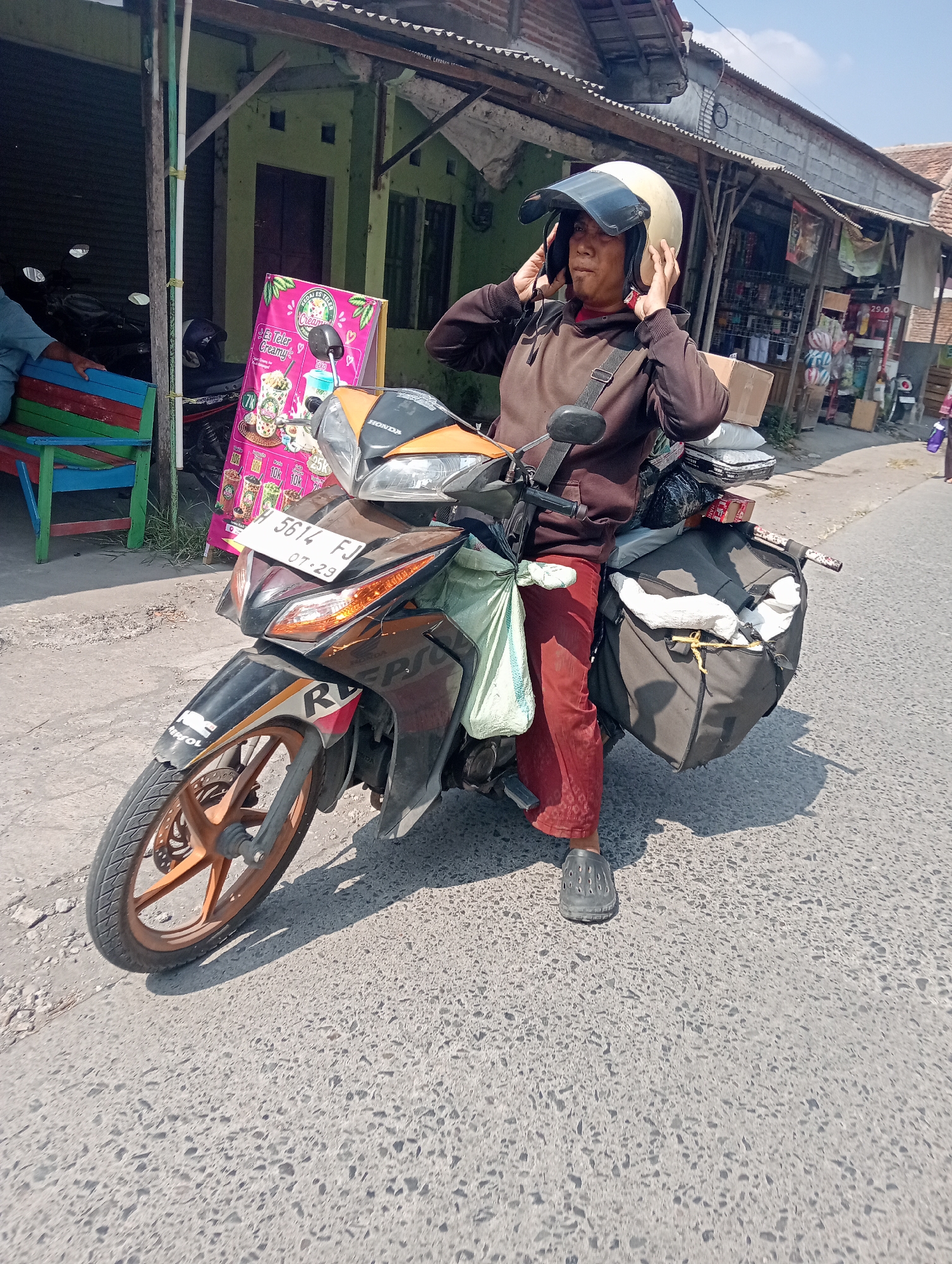 In the city of Katonsari, Indonesia, a man is shown adjusting his light-colored helmet while seated on a black and orange Honda motorcycle. The motorcycle is equipped with side bags, one of which is dark gray and filled with white and dark fabric, secured with yellow cord. Another light green bag is attached to the front of the motorcycle. The man is wearing a brown hooded sweatshirt and red pants, with dark gray clogs on his feet. The motorcycle has a license plate reading "H 5614 FJ" and "07-23". Behind the motorcycle, a vibrant pink advertisement displays text and images of drinks. The scene takes place on a paved road next to a building with a corrugated metal roof. Several other motorcycles are parked in the background, along with a multi-colored bench. The overall environment suggests a commercial or roadside setting under a clear sky.