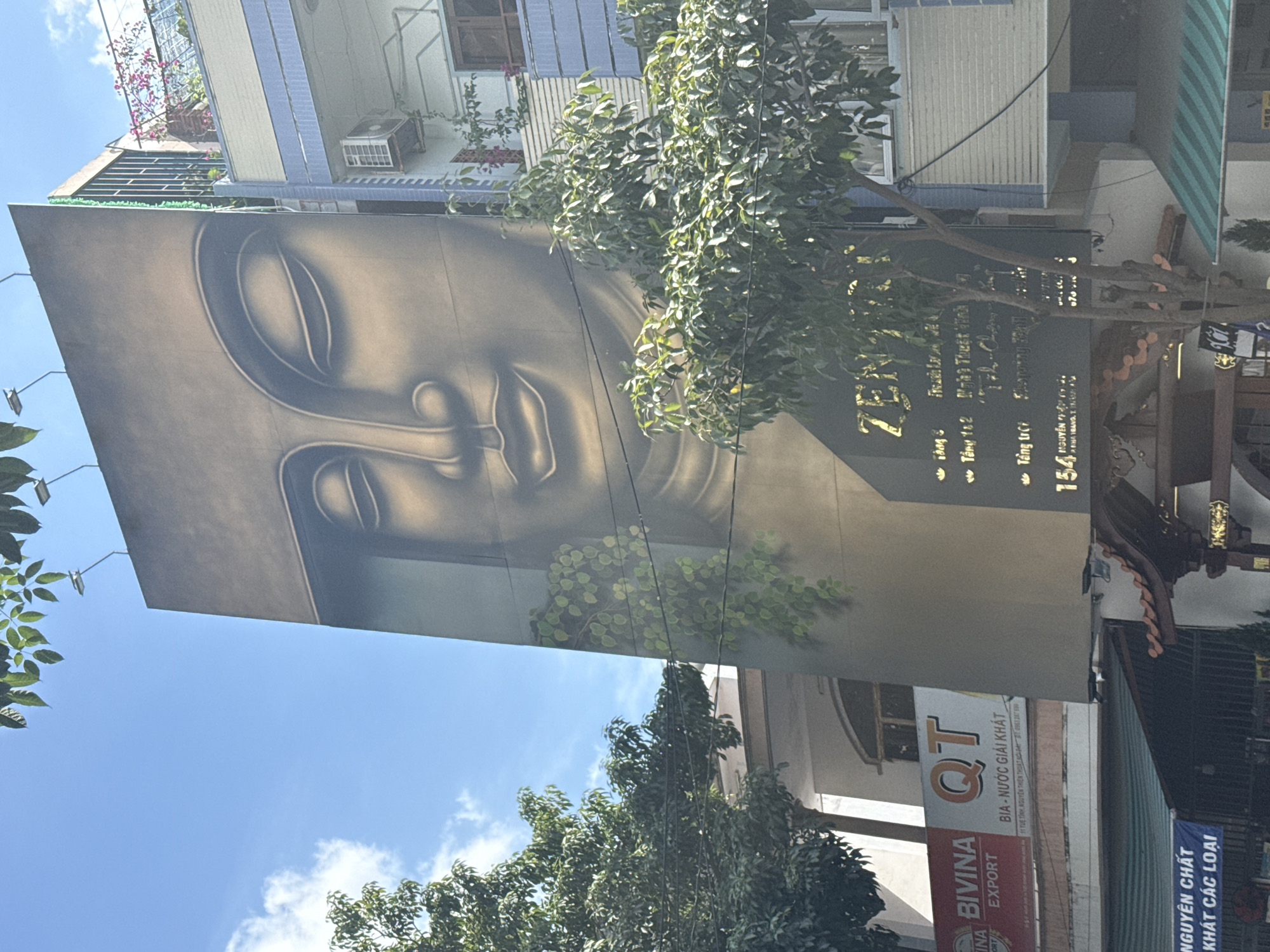 The image shows a tall building in Nha Trang Ward, Vietnam, with a large mural of a Buddha face on its side. The building also features a sign for "ZEN" with Vietnamese text, indicating it may be a spa or wellness center. The number 154 is also visible. Below ZEN are the words "Tanh Tri-Cho" and "skypaằng dahil". A Bivina Export sign for QT Bia-Nuoc Giai Khat (Beer-Drink for Thirst) is also visible on a sign for "Nguyen Chat Khat cac loai." Some trees partially obscure the view of the buildings. The sky is visible as well.