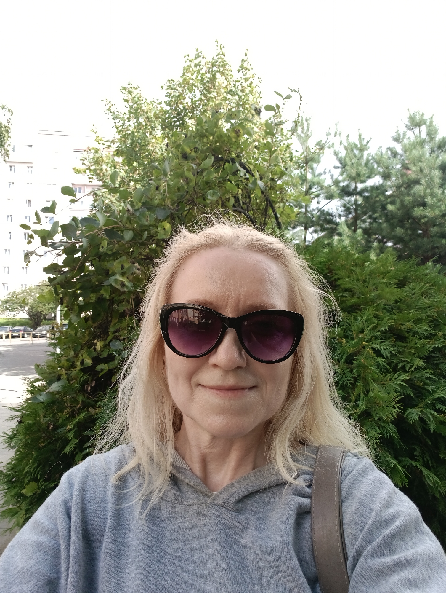 An outdoor, waist-up, medium-shot selfie shows a blonde woman with fair skin, wearing large, black, cat-eye sunglasses with purple lenses, and a light blue hooded sweatshirt. She has a slight smile. She is standing in front of lush green foliage. Behind the foliage, a tall white building with multiple windows and a street with parked cars are visible. The overall lighting suggests it is daytime. The woman is on a street in the city of Hrodna, Belarus.