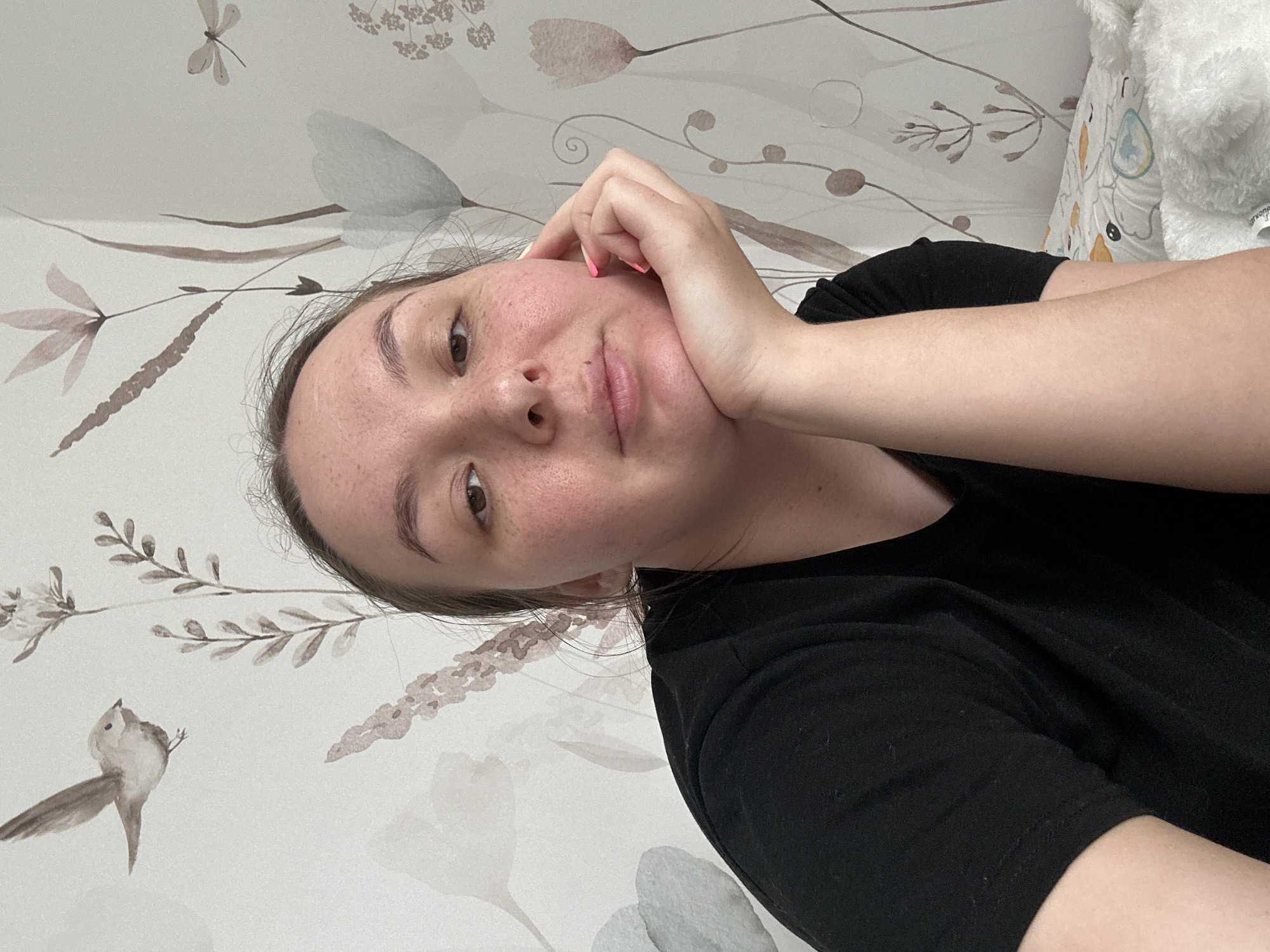 An image, oriented vertically, captures a young woman resting her head on her hand, looking towards the viewer. She appears to be lying down, with a relaxed and contemplative expression, exhibiting a subtle smile. Her face is visible with freckles across her nose and cheeks, and she has dark eyes and light brown hair. She is wearing a black short-sleeved t-shirt. Her right hand, supporting her head, shows pink nail polish.
The background features a light-colored wall or wallpaper adorned with muted, watercolor-style botanical illustrations including leaves, branches, and flowers in shades of brown, grey, and beige. Small animal figures, specifically a dragonfly and a bird, are also part of the pattern. To the right of the woman, a white, fluffy object, likely a blanket or pillow, is visible, partially obscuring a faint pattern and some Cyrillic text that appears to read "гкопа".
The scene suggests an indoor setting, possibly a bedroom or living space, in Chaikovsky, Russia, given the location context. The lighting is soft and even, implying daytime or a well-lit interior. There are no direct cues about the weather.