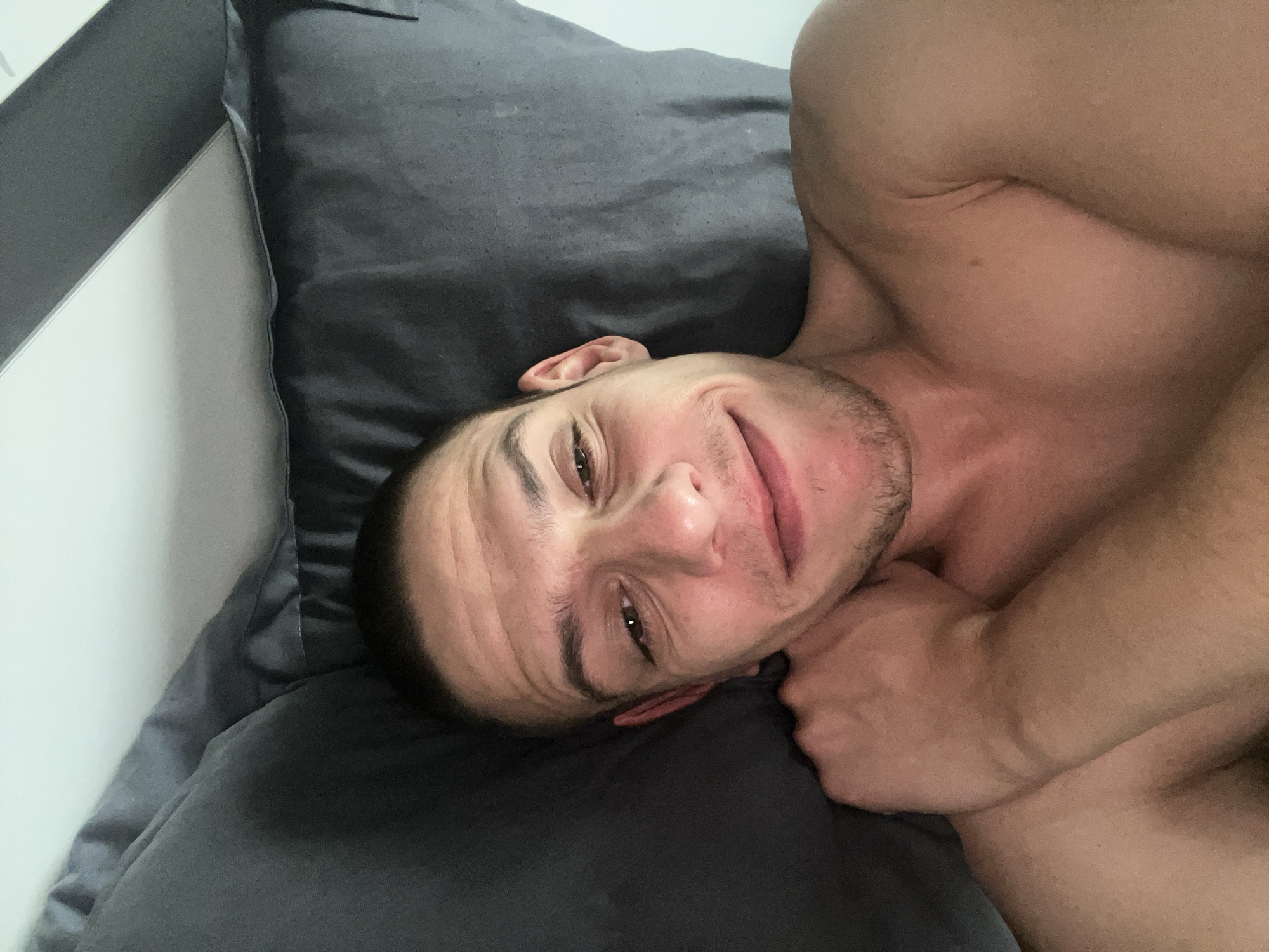 An eye-level shot captures a young adult male lying on his back, looking directly at the camera. He is shirtless, revealing a muscular physique, and is resting on a dark grey pillow. His head is turned slightly to face the viewer, and he has short, dark hair and dark eyes. A light stubble is visible on his face, which holds a relaxed, pleasant smile with slightly squinted eyes, conveying a friendly and calm demeanor. His left arm is partially visible, bent at the elbow, with his hand resting near his chest.
The setting appears to be indoors, likely a bedroom, characterized by the dark grey bedding that covers the bed. A crisp white wall or headboard is visible along the left edge of the frame, providing a contrasting backdrop. The lighting is soft and natural-looking, illuminating his features without harsh shadows. No text or specific time of day is discernible from the image.