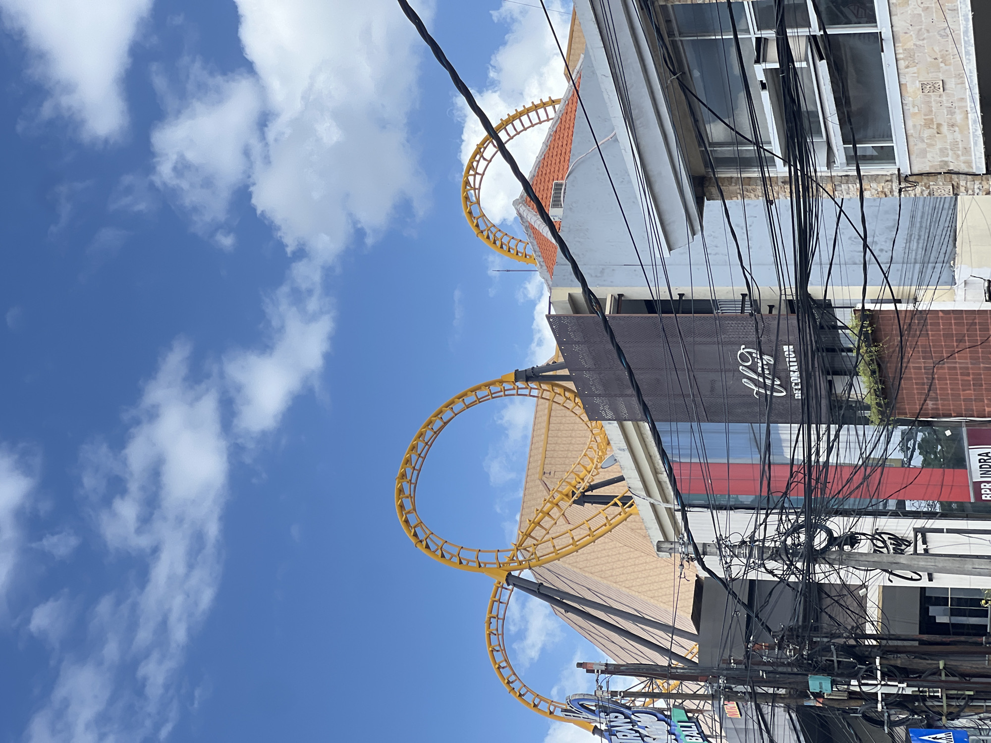 The image captures a street scene in Denpasar, Indonesia, dominated by a bright blue sky with scattered white clouds. In the background, a yellow roller coaster track with two loops is prominently visible, seemingly integrated with or adjacent to a building with a tan, angled roof. The building beneath the roller coaster has a multi-story facade featuring glass windows and sections with red and black accents. A blue sign with white lettering, partially obscured by electrical wires, reads "TRANS" and "BALI".
In the foreground, a dense network of black electrical wires hangs across the frame, supported by utility poles. These wires create a complex, tangled web that partially obscures the view of the buildings behind them. Several buildings line the street, exhibiting various architectural styles and materials, including glass, concrete, and brick. One building on the right features a light-colored stone facade with several windows, some of which are open. Another building on the left has a dark facade with text that includes the words "DECORATION" in white lettering. A street sign with a pedestrian crossing symbol is visible at the bottom left of the image. No people are clearly discernible in the image. The overall impression is one of a busy urban environment with a blend of modern structures and functional infrastructure, under a clear sky.