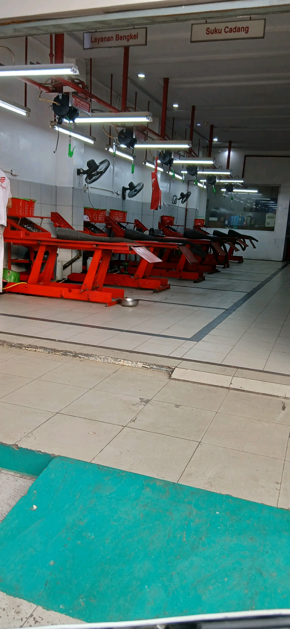 The image shows the interior of a motorcycle repair shop in Samarinda, Indonesia. The shop features multiple red motorcycle lifts arranged in parallel. Overhead, fluorescent lights illuminate the space, and signs are visible reading "Layanan Bengkel" (Workshop Services) and "Suku Cadang" (Spare Parts). Several black oscillating fans are mounted on the walls. In the foreground, a teal-colored ramp is present on the tiled floor. The overall environment suggests a functional and organized workshop space.