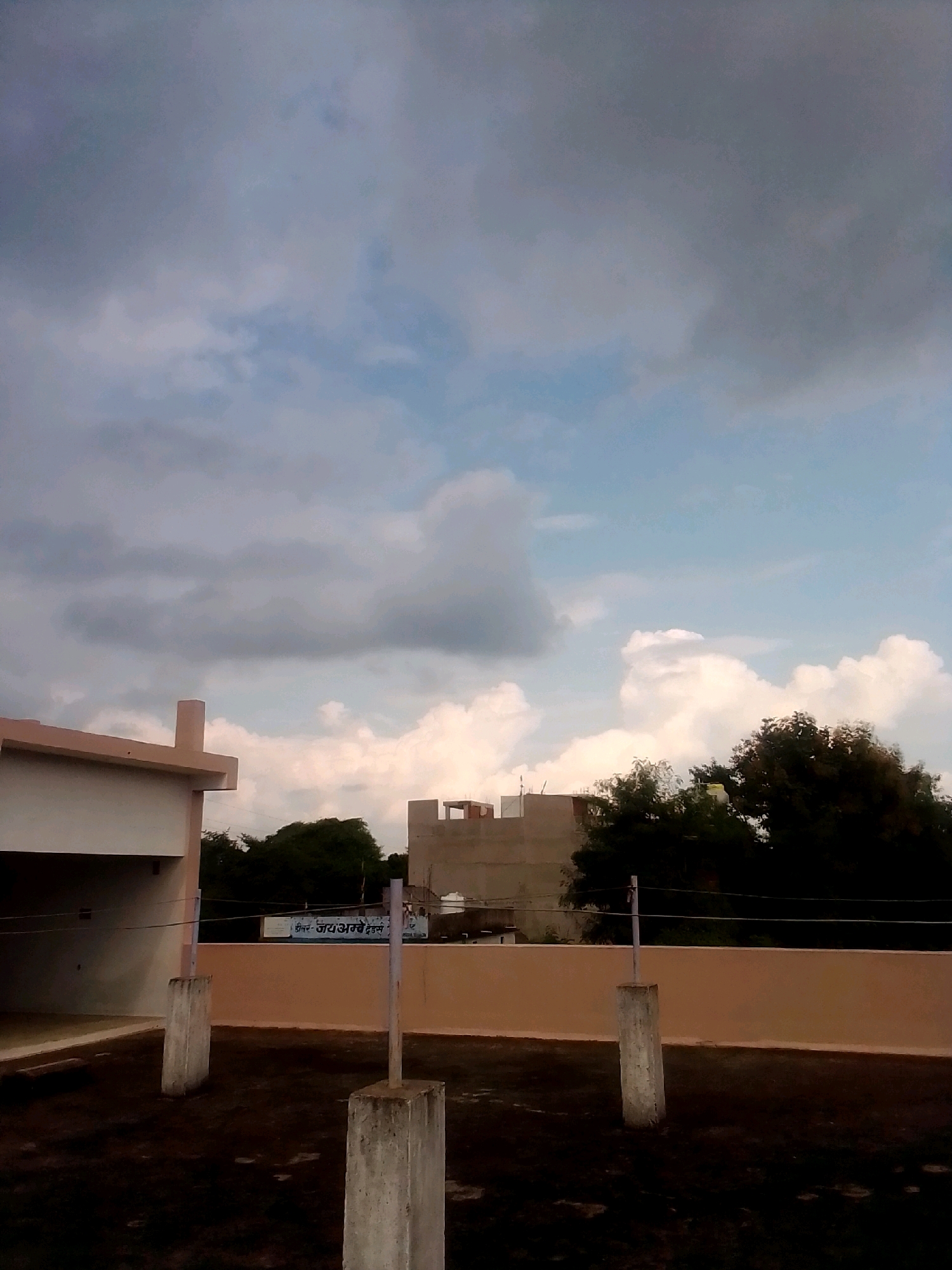 The image depicts a rooftop view in Anjaniya, India, under a cloudy sky. In the foreground, a dark, wet-looking rooftop is visible, with two concrete pillars supporting thin wires stretched across. To the left, a portion of a light-colored building with a beige overhang is present. In the background, several buildings of varying heights are seen, some with visible construction. Notably, a billboard with text that appears to be in Hindi is attached to a wall. Lush green trees form a dense line behind the buildings. The overall atmosphere suggests an overcast day, possibly before or after rain.