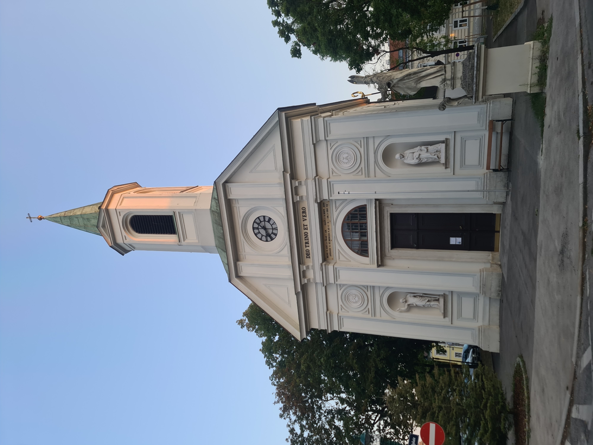 This is a low-angle, eye-level, medium shot of the exterior of a white chapel in Vienna, Austria. The chapel has a steeple topped with a cross, a clock on its facade, and two statues in niches on either side of the entrance. The entrance has a dark, wooden double door. A statue stands to the right of the chapel. The sky is clear and blue, with the setting sun casting a warm glow on the steeple. Trees and a street sign are visible in the background.