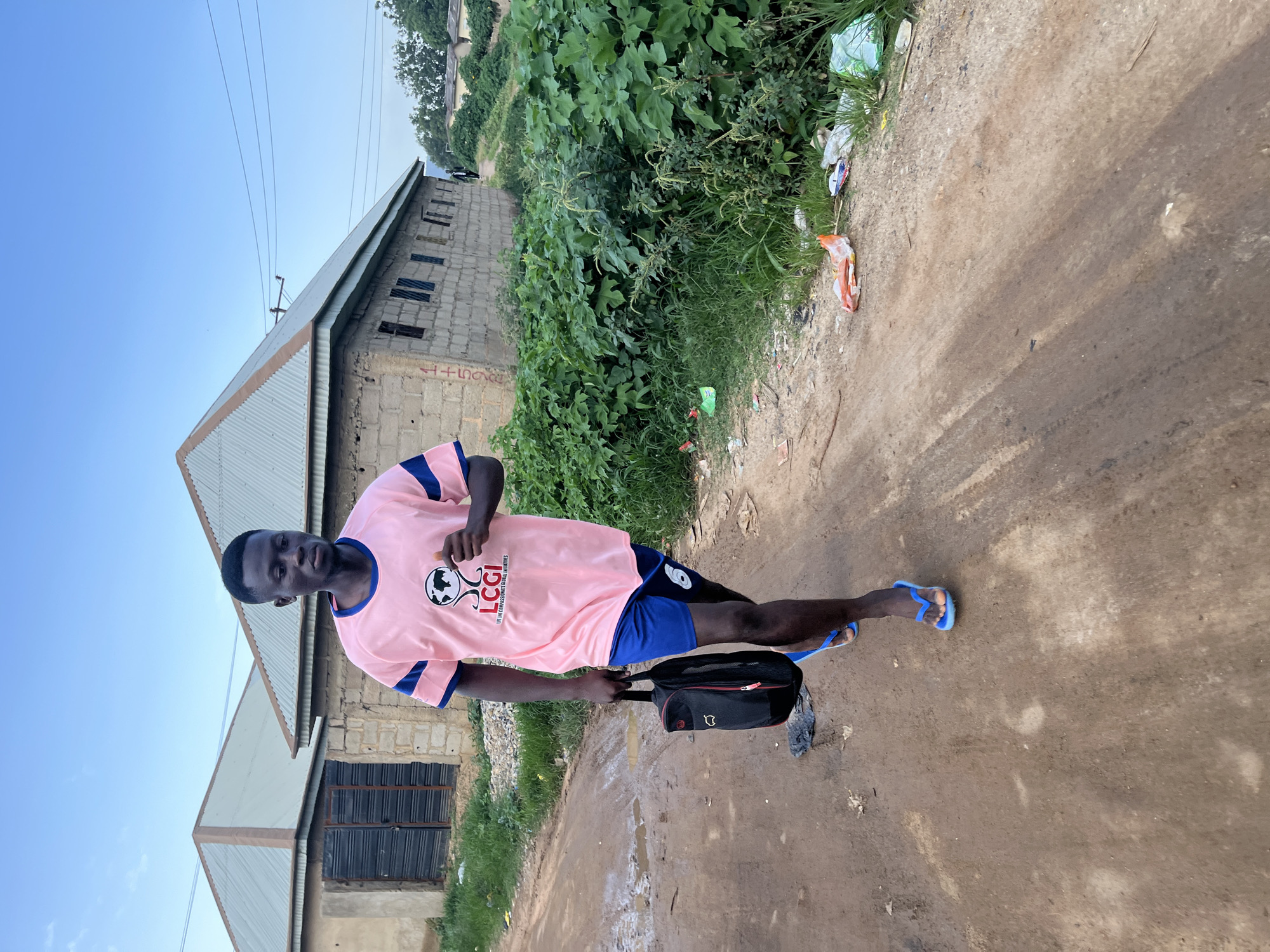 A young man is walking down a dirt road in Rafin Par, Nigeria. He is wearing a pink t-shirt with a graphic on it and blue shorts. He is carrying a black bag in his left hand and wearing blue flip-flops. The road is unpaved and appears to be wet in places, suggesting it may have rained recently. There are buildings on either side of the road, constructed from concrete blocks. Lush green vegetation lines the road and the surrounding area, with some scattered litter visible on the side of the road. The sky is a clear blue with some scattered clouds. The man is walking away from the camera, but his face is partially visible and he appears to be looking forward.