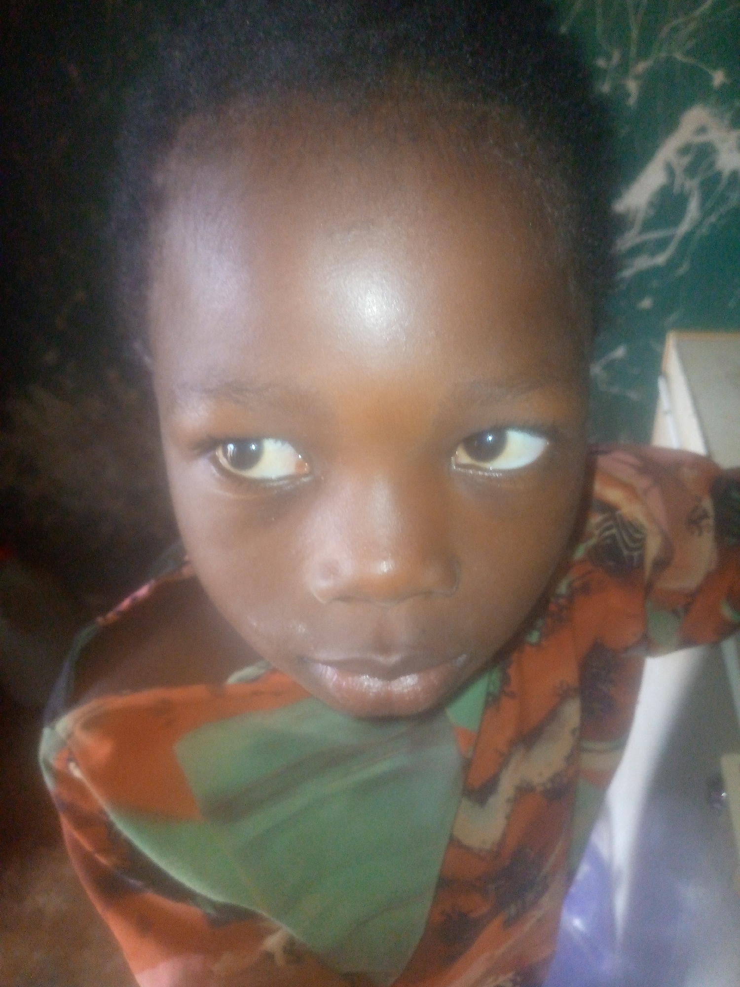 The image features a young child, likely a toddler or in early childhood, looking directly at the camera. The child has dark skin and short, dark, curly hair. Their eyes are wide open with dark brown irises, and a highlight suggests a light source reflecting in them. The camera is positioned high, looking down at the child from above.
The child is wearing a garment that has a prominent green section and patches of orange and brown with darker, abstract patterns. The fabric appears soft and draped.
The background is mostly dark and out of focus. To the child's left (viewer's right), a greenish surface with distinct white, swirly patterns is visible, possibly a patterned wall or a large piece of fabric. Further to the right, a lighter, off-white or beige object, possibly a piece of furniture or a door frame, can be discerned. The overall lighting suggests an indoor setting, possibly with artificial light, given the highlights on the child's skin and the darkness of the background.
The child's expression is neutral and direct, conveying a sense of curiosity or observation. No specific activity is taking place other than the child looking at the camera. There is no visible text in the image.