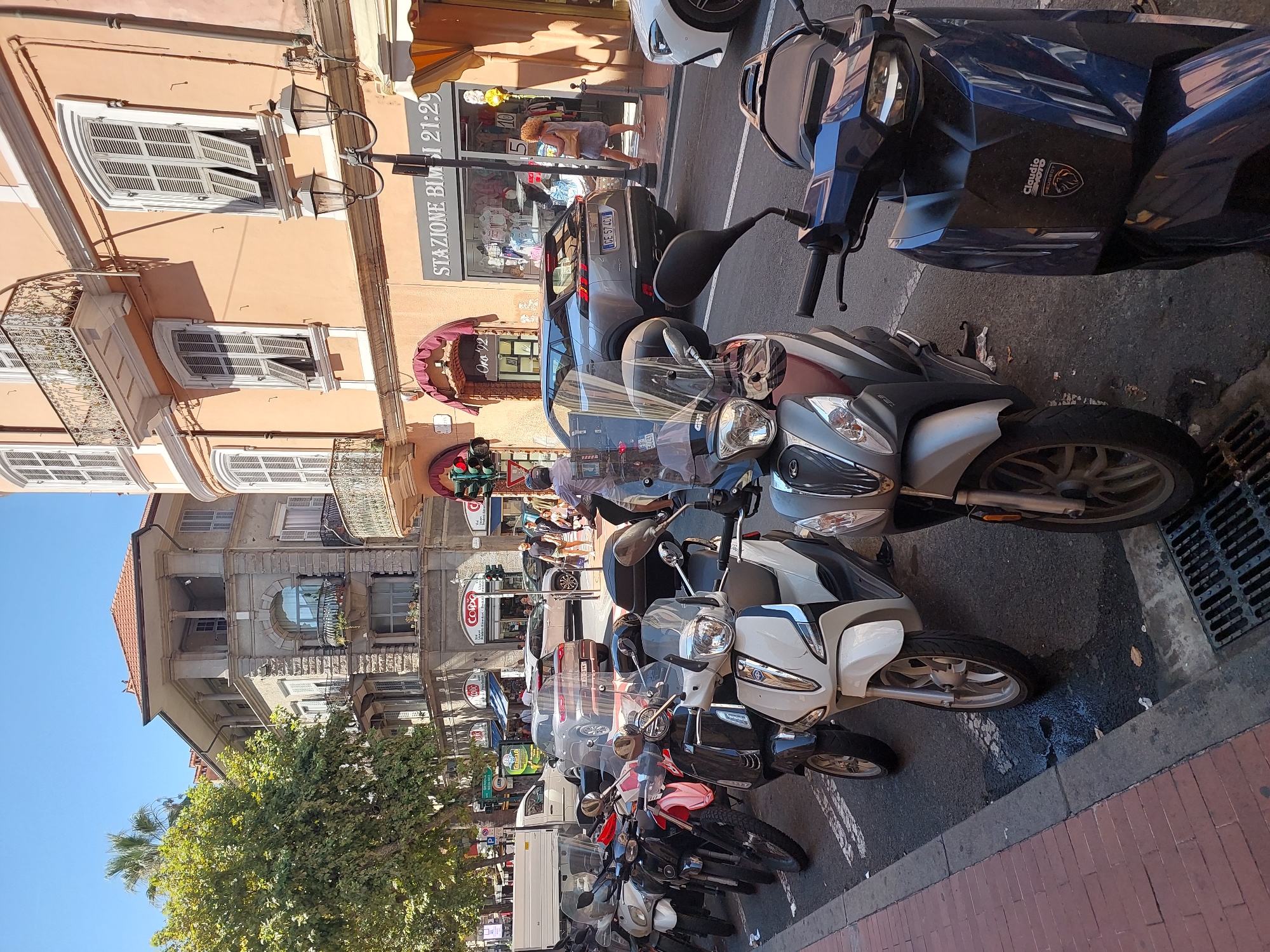 The image depicts a street scene in Ventimiglia, Italy, on a sunny day. In the foreground, a row of parked scooters lines the sidewalk. The scooters are of various colors, including white, black, red, and blue. Some of the scooters have windshields and storage boxes. One blue scooter in the far right foreground has a logo that appears to be "CLAUDIO MOTO PEUGEOT".
Behind the scooters, the street is visible, with cars and pedestrians. A silver SUV is parked on the street, and a white van is also present. People are walking on the sidewalk and crossing the street. The buildings lining the street are old and have an Italian architectural style, with balconies and shuttered windows. One building is painted a pale orange, and another is a lighter stone color. Signs for businesses are visible, including one that reads "STAZIONE BIMBI 21:29". Traffic lights are also present at an intersection.
The overall impression is a typical European street scene on a warm day, with a mix of vehicles and pedestrians. The presence of scooters suggests a popular mode of transportation in the area. The architecture and signage indicate a location in Italy.