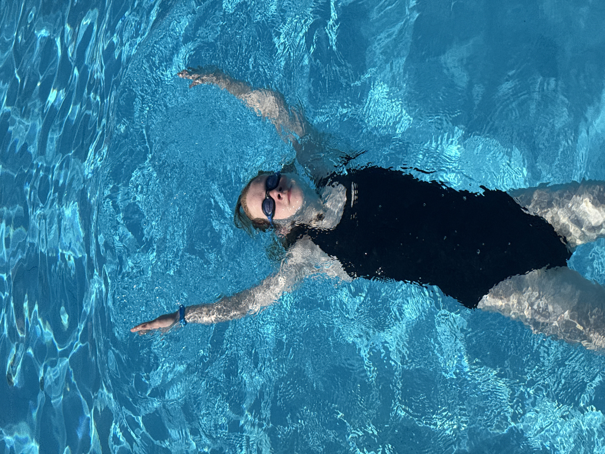 The image displays a young individual, possibly female, floating supine in a clear, blue swimming pool in Nebug, Russia. The individual is wearing a black one-piece swimsuit and blue swimming goggles. Her arms are extended upwards and partially submerged, with a blue wristband visible on her left wrist. Her head is tilted slightly back, and her eyes appear closed or directed towards the sky, indicating a state of relaxation. The pool water exhibits strong light refractions and ripples across its surface, consistent with an outdoor, sunlit environment. Dark edges of the pool are discernible in the lower and right portions of the frame.