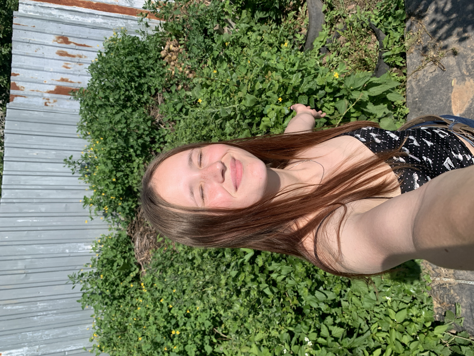 In a selfie taken outdoors, a young Caucasian female with long, brown hair and closed eyes smiles, her face illuminated by sunlight. She is wearing a black and white polka dot camisole. Her left arm is extended, holding the camera. The background consists of dense green foliage with scattered yellow flowers, and a weathered, corrugated metal fence. The setting appears to be a rural or semi-rural area, possibly within the city of Foky, Russia, given the natural vegetation and fencing. The overall impression is one of casual enjoyment on a sunny day.
