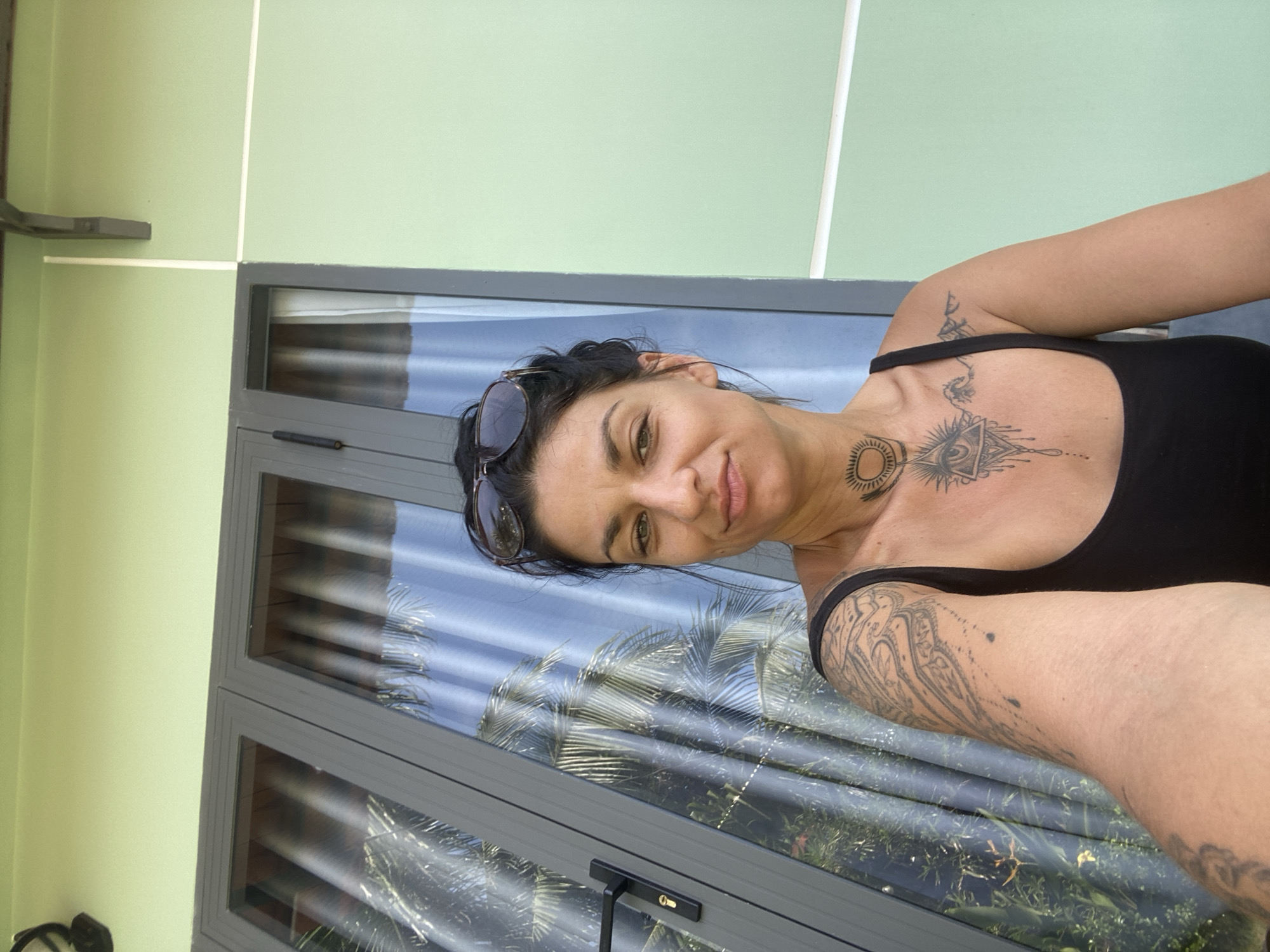 In Mũi Né, Vietnam, a woman with dark hair pulled back and sunglasses perched on her head, is captured in a selfie. She wears a black tank top, revealing tattoos on her arm, chest, and neck. The background features a light green building with large glass doors reflecting palm trees and the sky. The image appears to have been taken outdoors, likely on a balcony or patio.